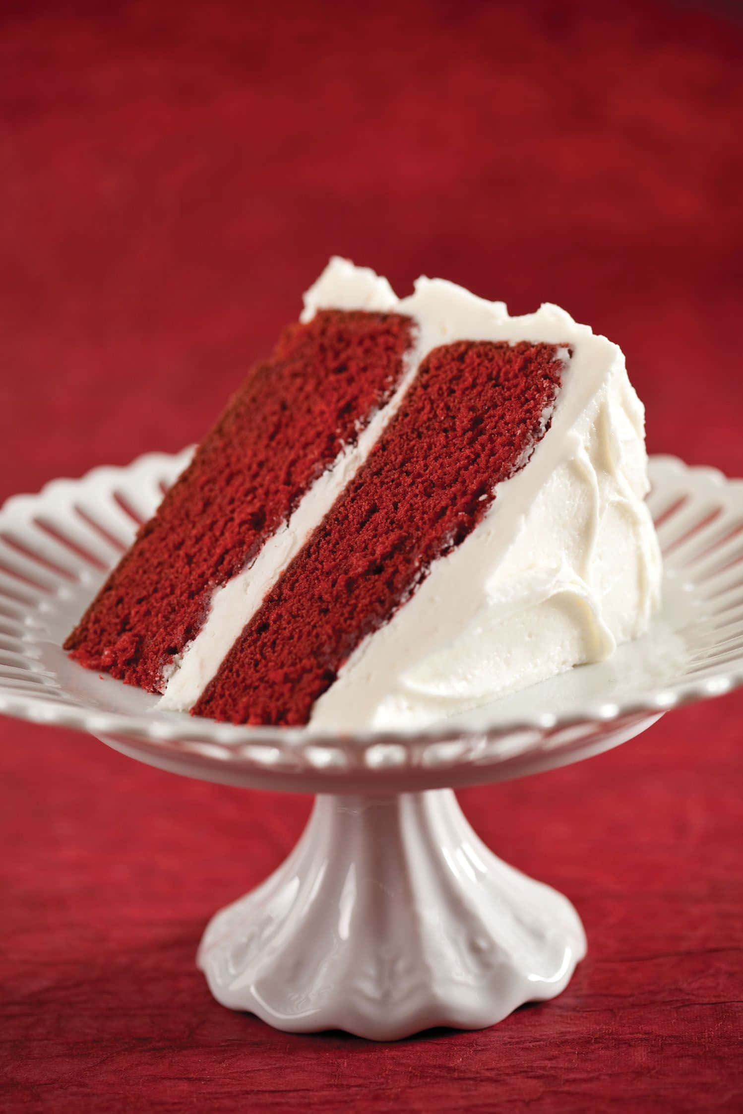Red Velvet Cake 桌布 壁紙 — 一塊頹廢的紅絲絨蛋糕，配上奶油白色糖霜，優雅地放在白色陶瓷底座上，背景是濃郁的紅色。