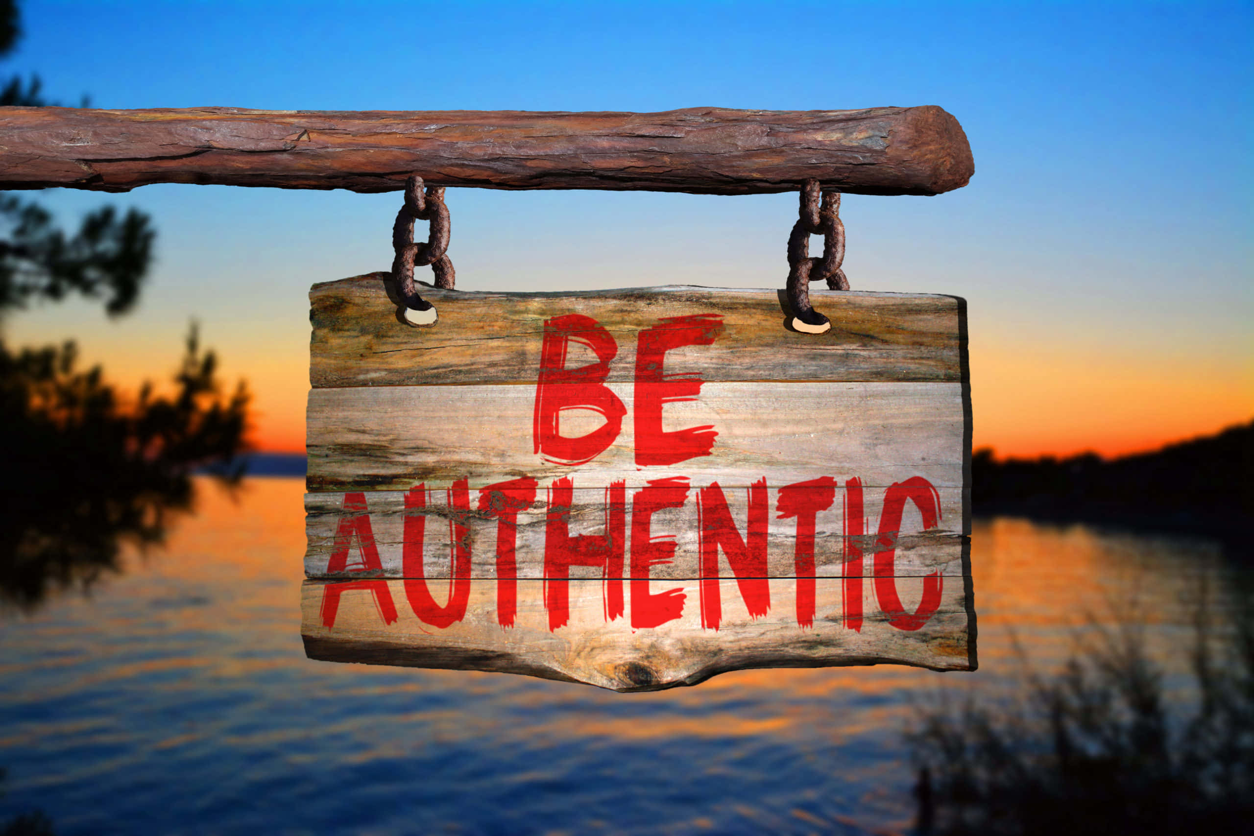 Authentic 桌布 壁紙 — 一塊飽經風霜的木牌，上面用粗體紅色字母寫著「BE AUTHENTIC」，懸掛在日落時分平靜的湖面上。
