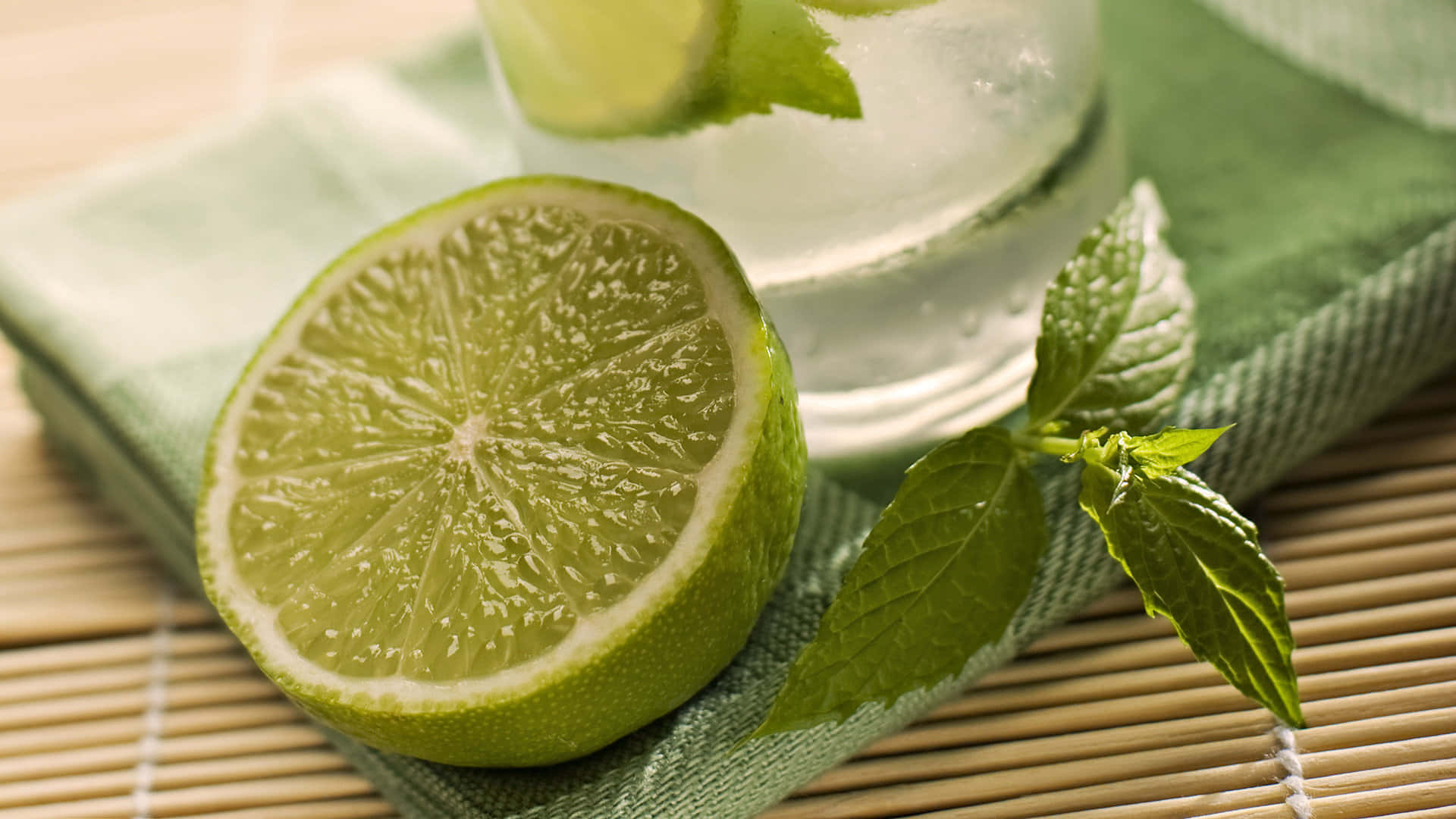 Lime 桌布 壁紙 — 一塊鮮豔的青檸半邊和新鮮薄荷葉放在竹上綠色布料的冰凍飲品杯旁。