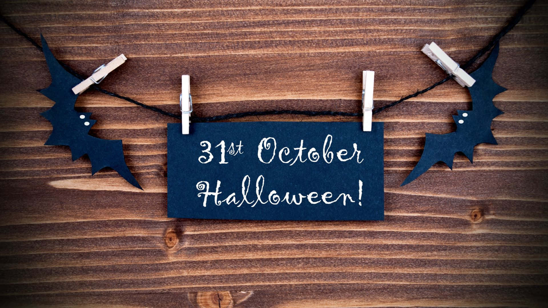 October 31st 桌布 壁紙 — 一塊黑板標誌寫著10月31日萬聖節，掛在一條黑色繩子上，帶有蝙蝠剪影和衣夾，背景是質樸的木質表面。