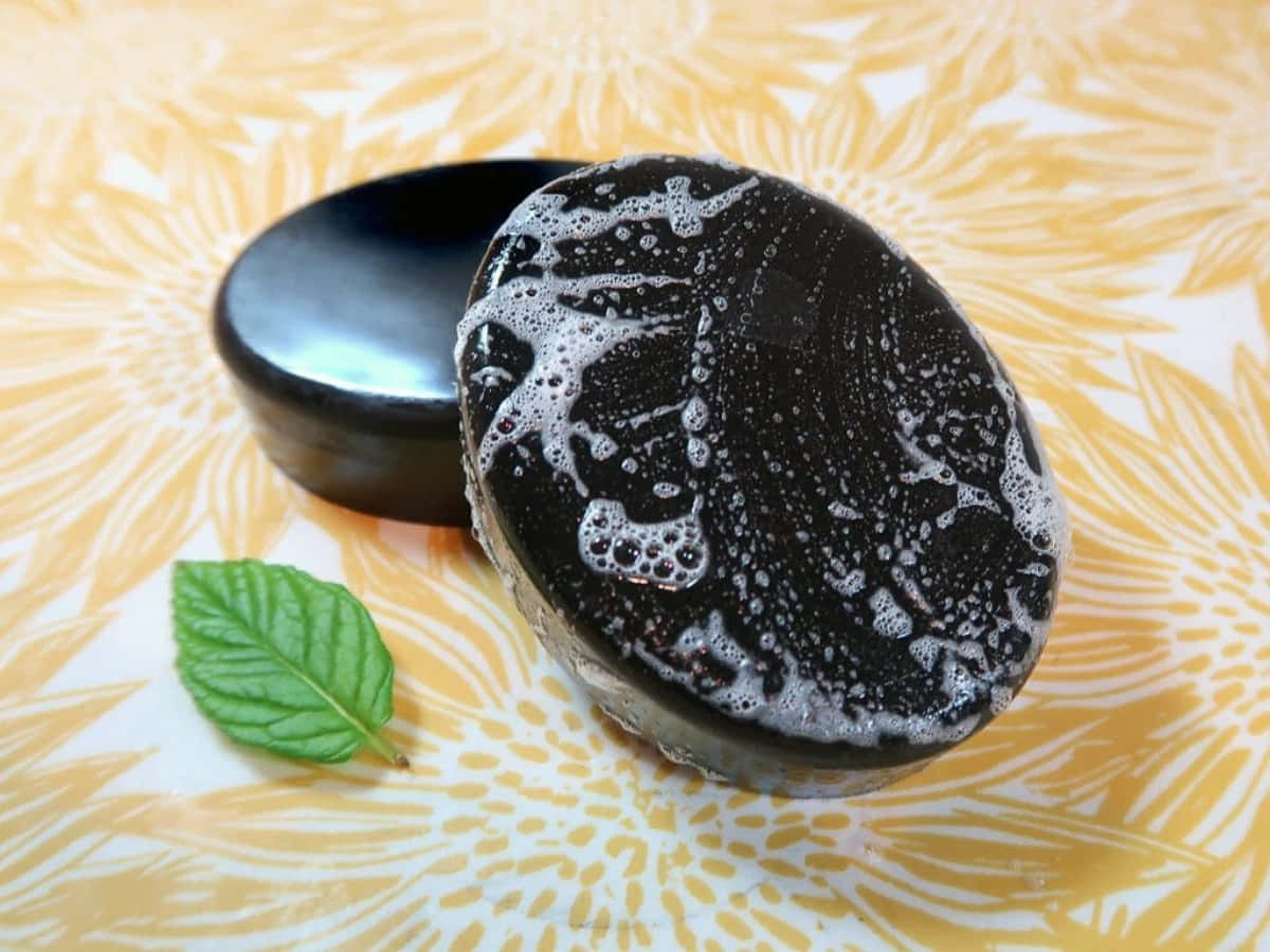 Black Soap 桌布 壁紙 — 一塊黑色肥皂帶有白色泡沫，旁邊放著蓋子和一片新鮮薄荷葉，置於黃色花卉圖案表面上。