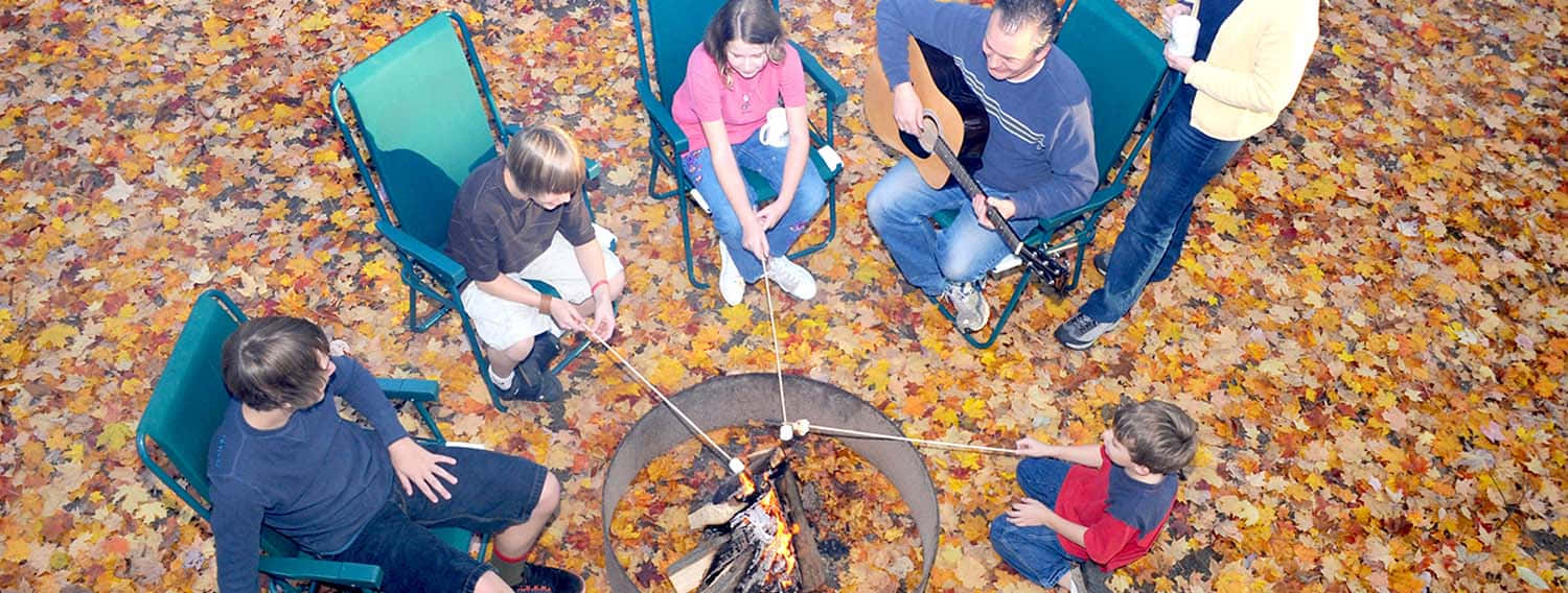 Fall Camping 桌布 壁紙 — 一家人圍著營火，用樹枝烤棉花糖，周圍鋪滿金黃色的秋葉。