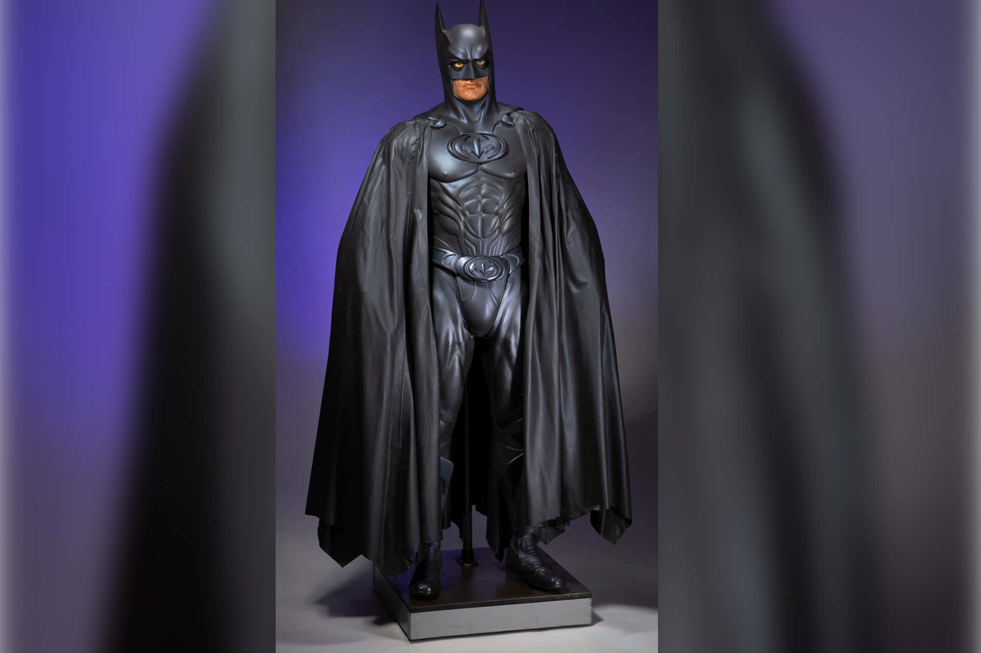 Bat-suit 桌布 壁紙 — 一尊亮黑色蝙蝠俠雕像矗立在基座上，背景為紫色漸變，帶有戲劇性的燈光效果。