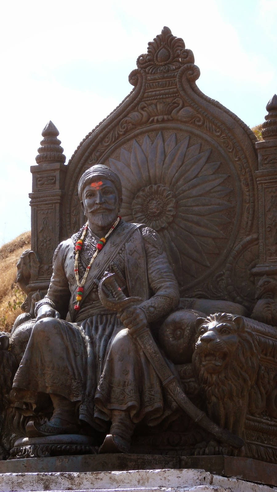 Indian 桌布 壁紙 — 一尊威嚴的青銅雕像，一位留鬍鬚的男子身著傳統服飾，端坐在裝飾華麗的寶座上，兩側有獅子雕塑，背景是明亮的天空。