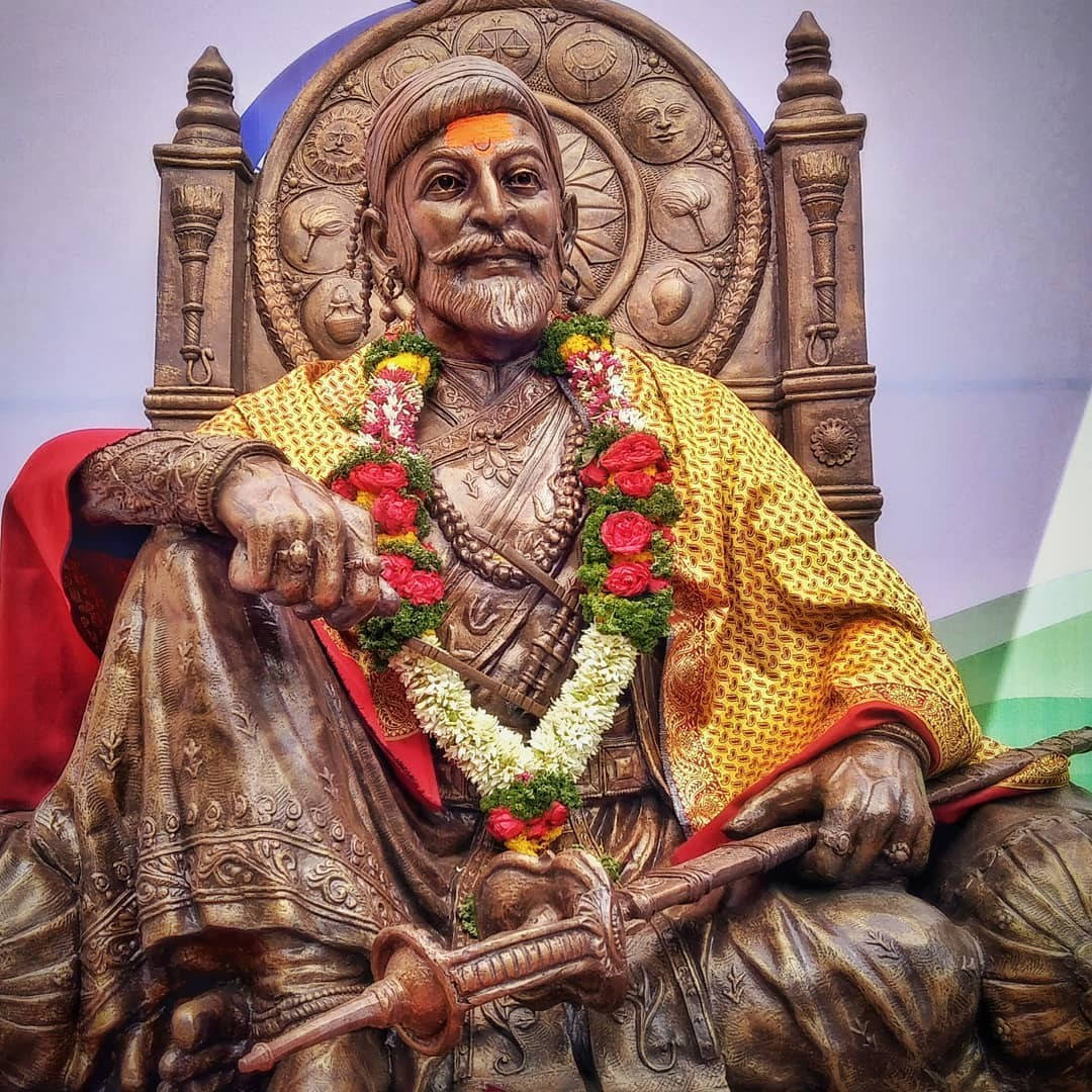 Indian 桌布 壁紙 — 一尊威嚴的鬍鬚國王青銅雕像，坐在裝飾著紅白花環的華麗王座上。