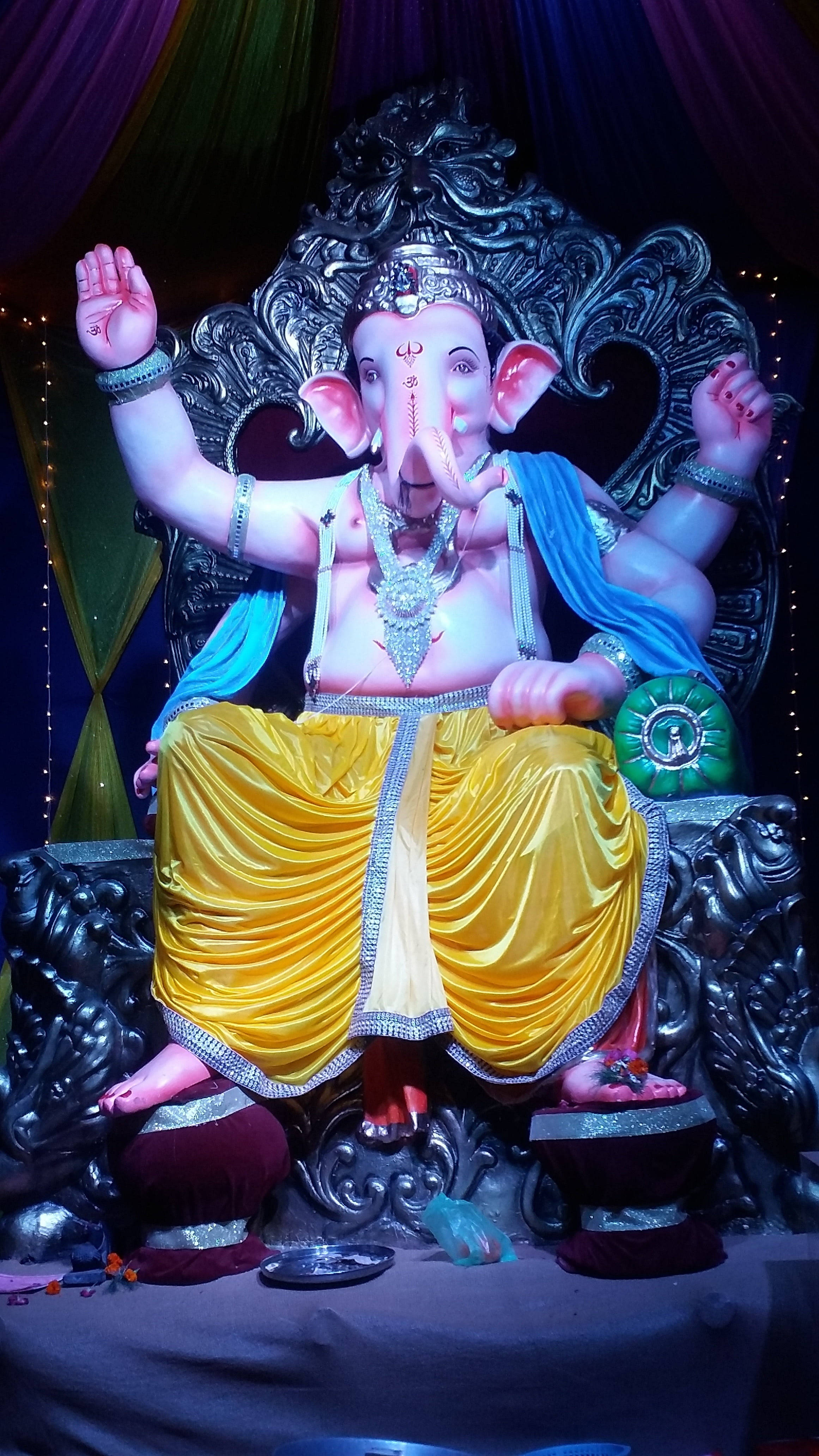 ganesh iPhone 桌布 壁紙 — 一尊宏偉的象神雕像威嚴地坐在華麗的寶座上，寶座覆蓋著金色和藍色布料，上方是色彩繽紛的垂簾。