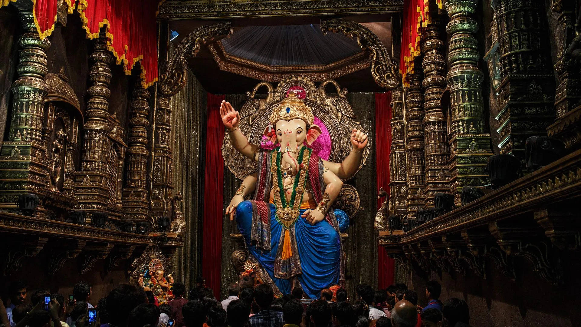 Hindu God 桌布 壁紙 — 一尊宏偉華麗的象頭神像身穿藍色長袍，威嚴地坐在裝飾華麗的舞台上，信徒們聚集在下方。