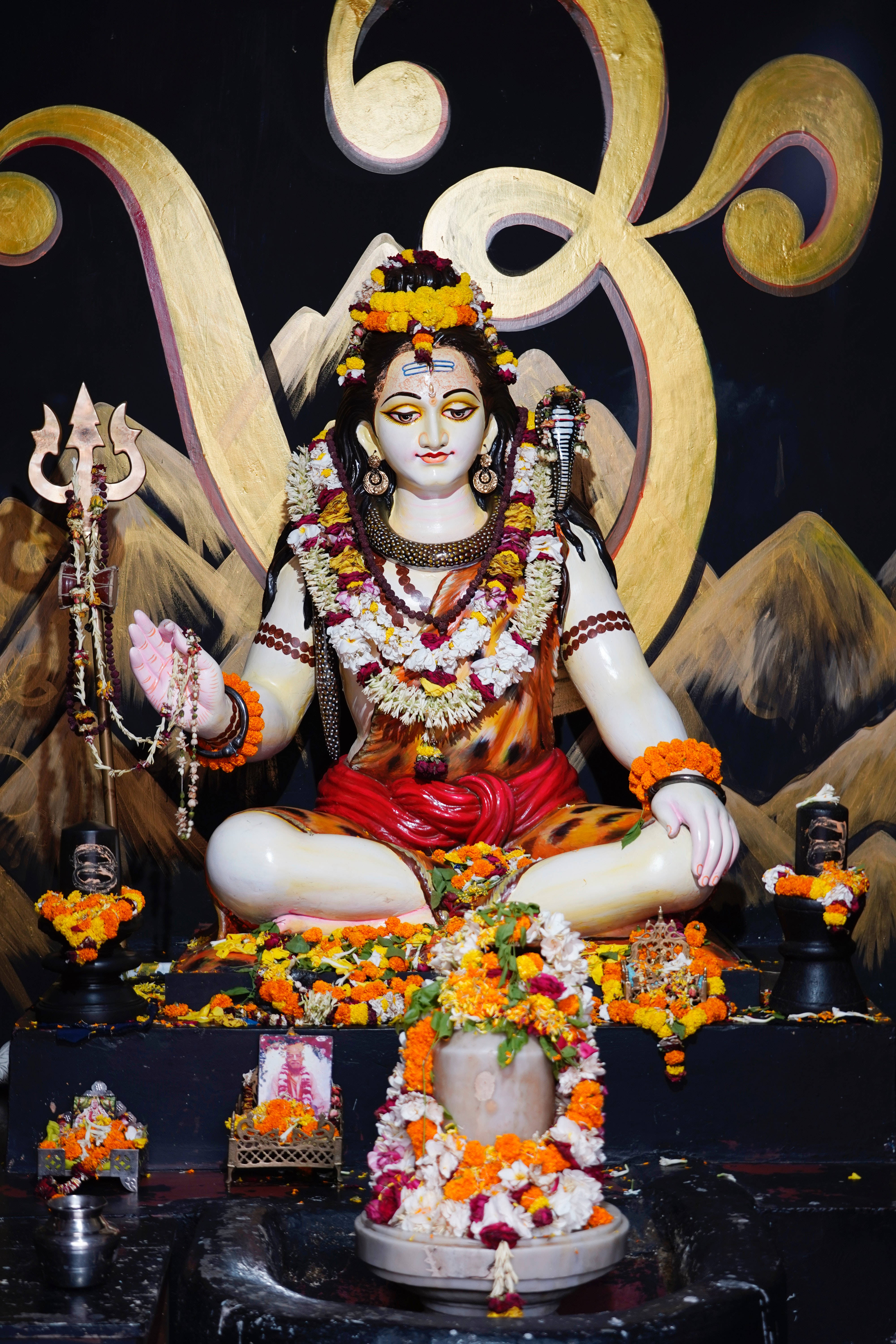 Lord Shiva 桌布 壁紙 — 一尊寧靜的濕婆神像，飾以鮮艷的橙色和白色萬壽菊花環，盤腿坐在裝飾華麗的祭壇上，背景是金色的華麗圖案。