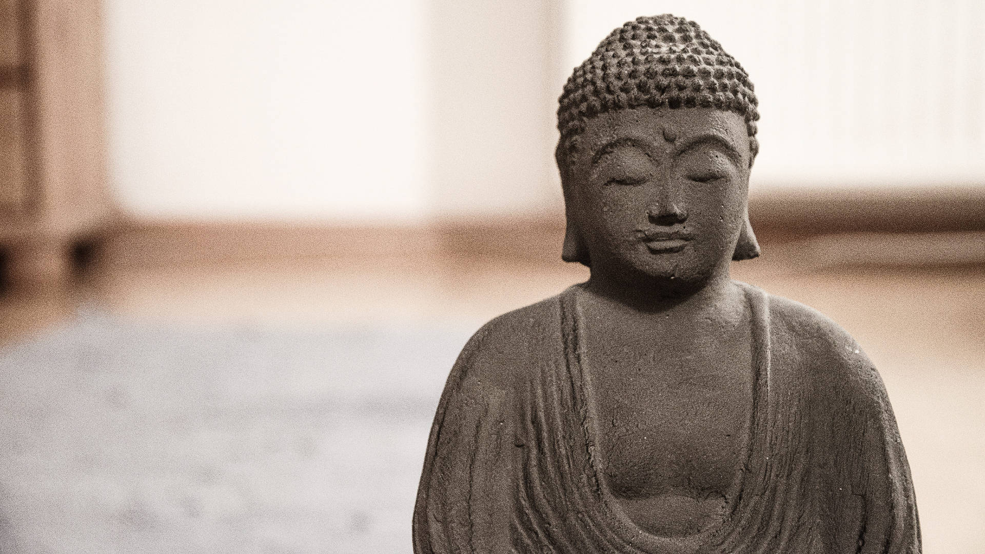 Buddha 桌布 壁紙 — 一尊寧靜的石佛像，閉著眼睛，穿著有紋理的長袍，在溫暖模糊的室內背景下以柔焦呈現。
