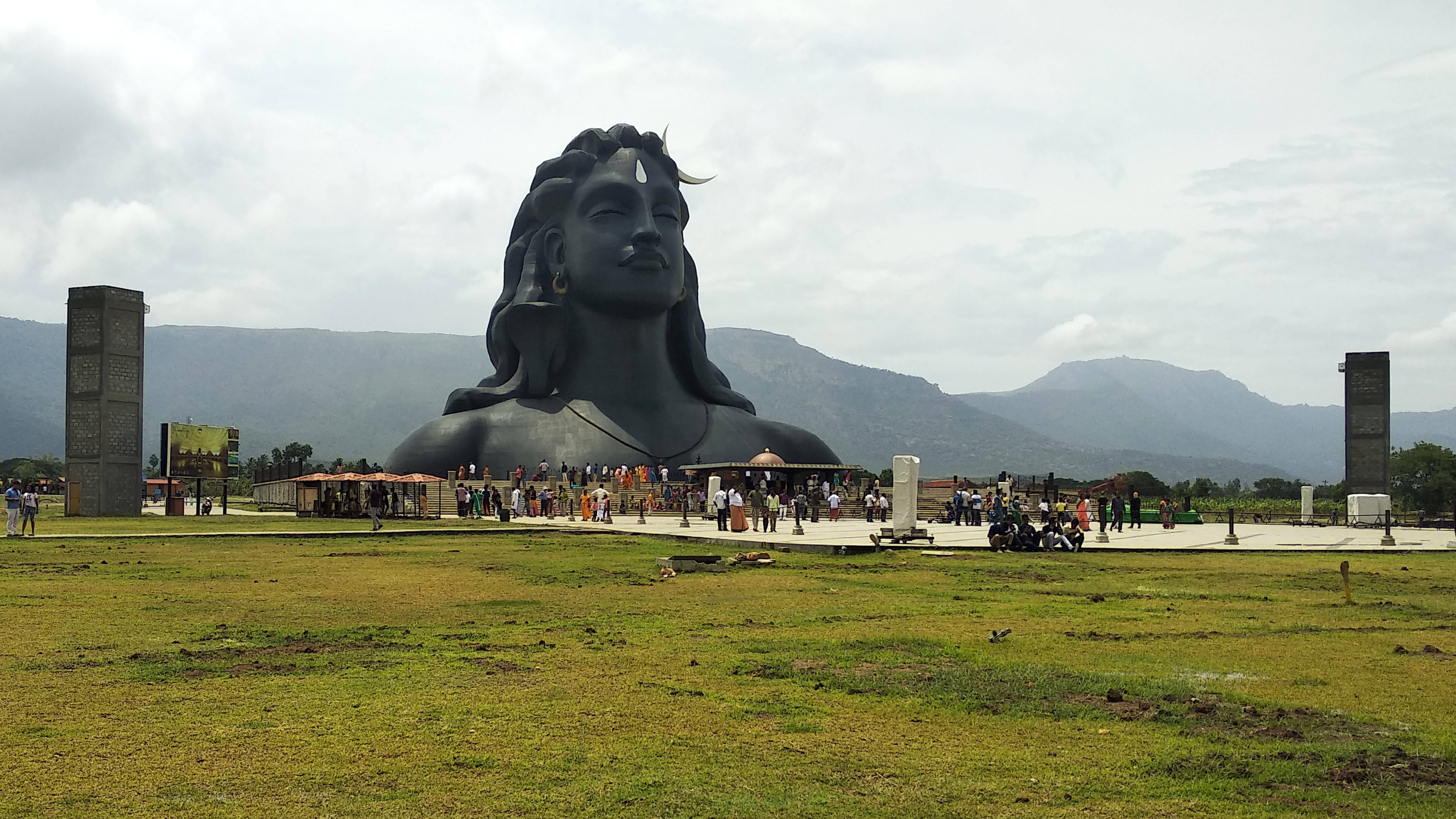Lord Shiva 桌布 壁紙 — 一尊巨大的深色石像，描繪帶有新月和第三隻眼的濕婆神，俯瞰著一片草地，草地上有遊客和遠處的山脈，天空多雲。