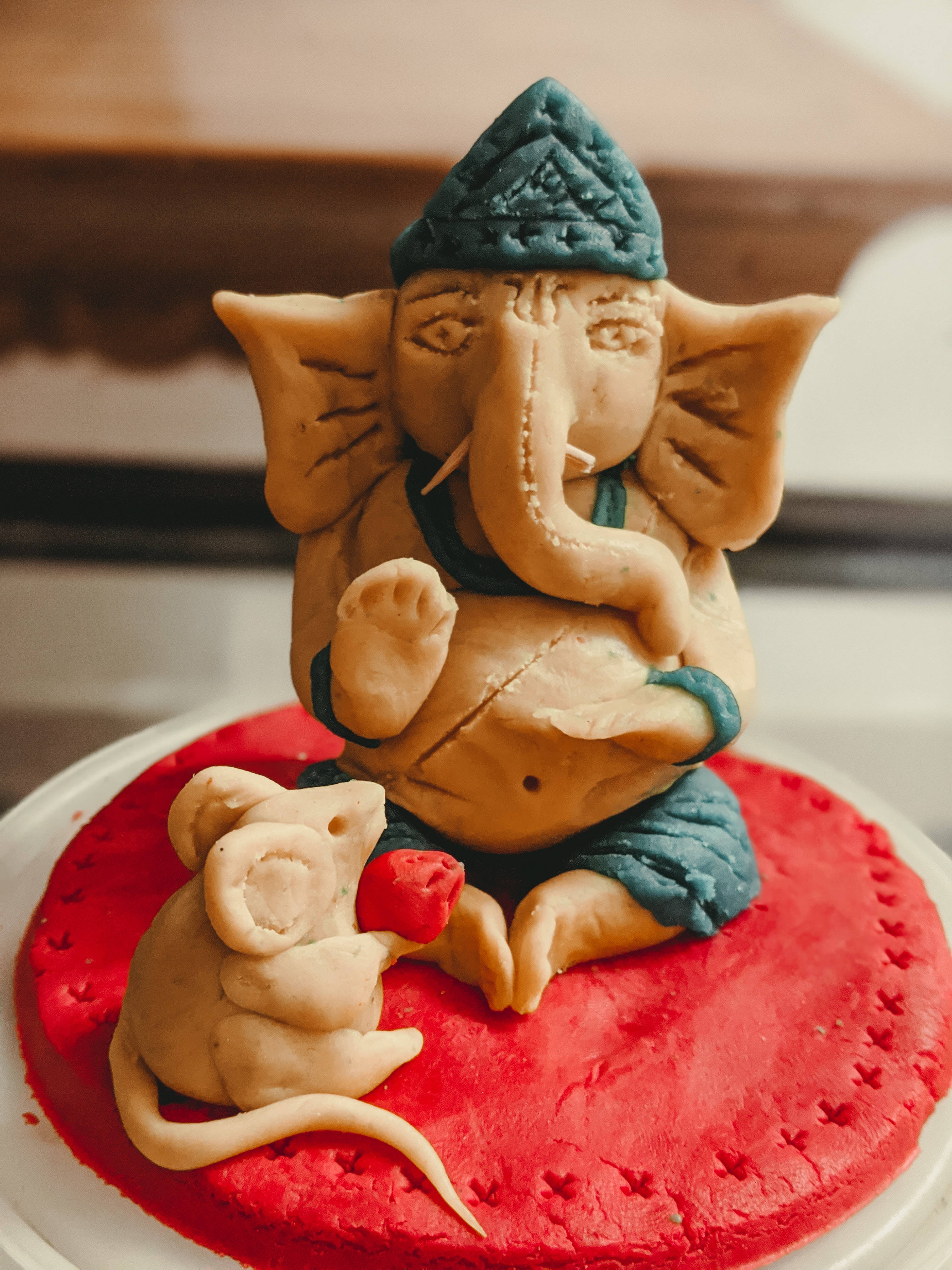 Ganesh 桌布 壁紙 — 一尊手工製作的象頭神甘尼許黏土雕像，伴隨著一隻老鼠，坐在紅色紋理底座上。