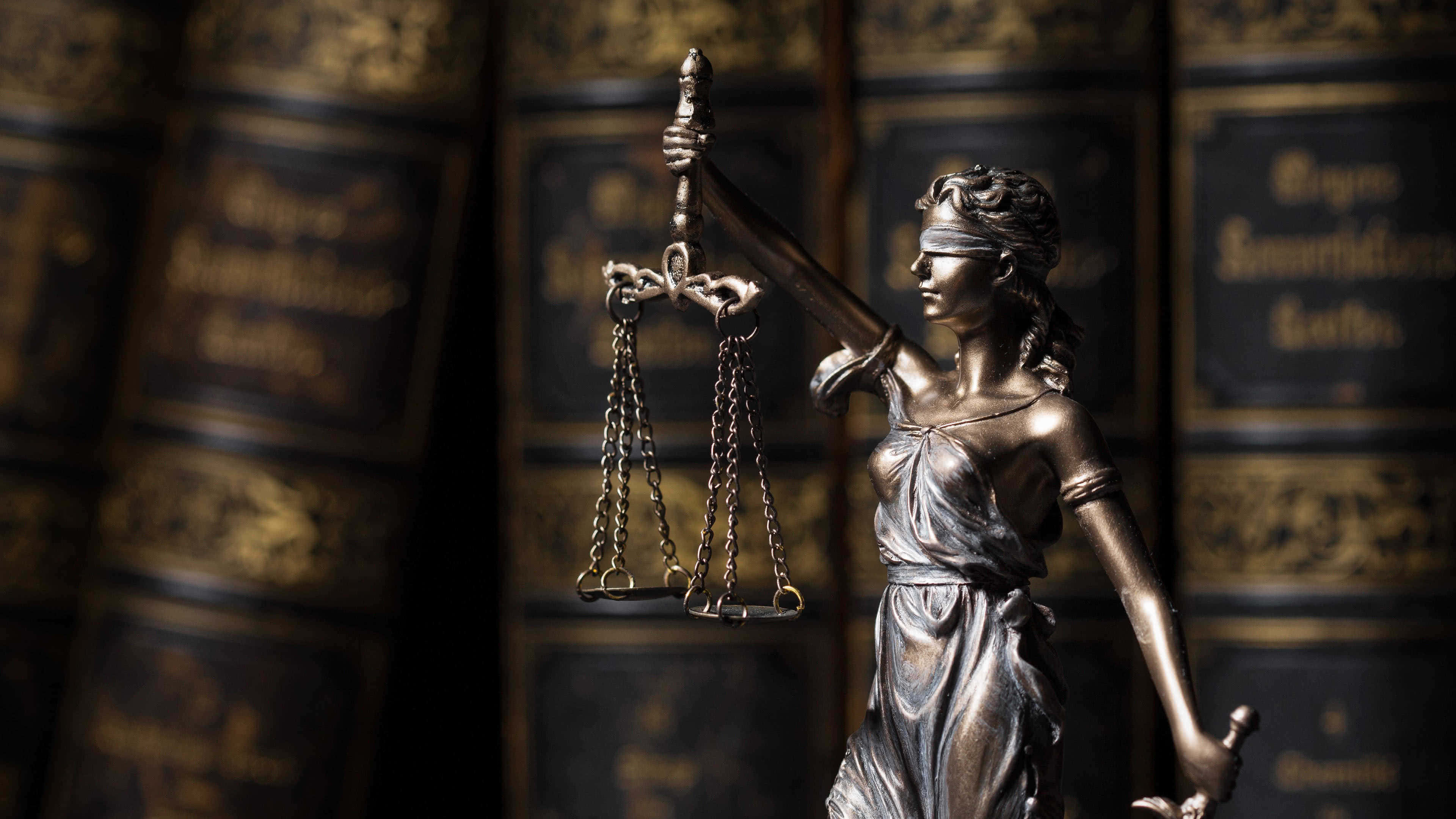 Justice 桌布 壁紙 — 一尊手持天秤和劍的正義女神青銅雕像，立於裝飾華麗的法律書籍前，環境燈光昏暗。