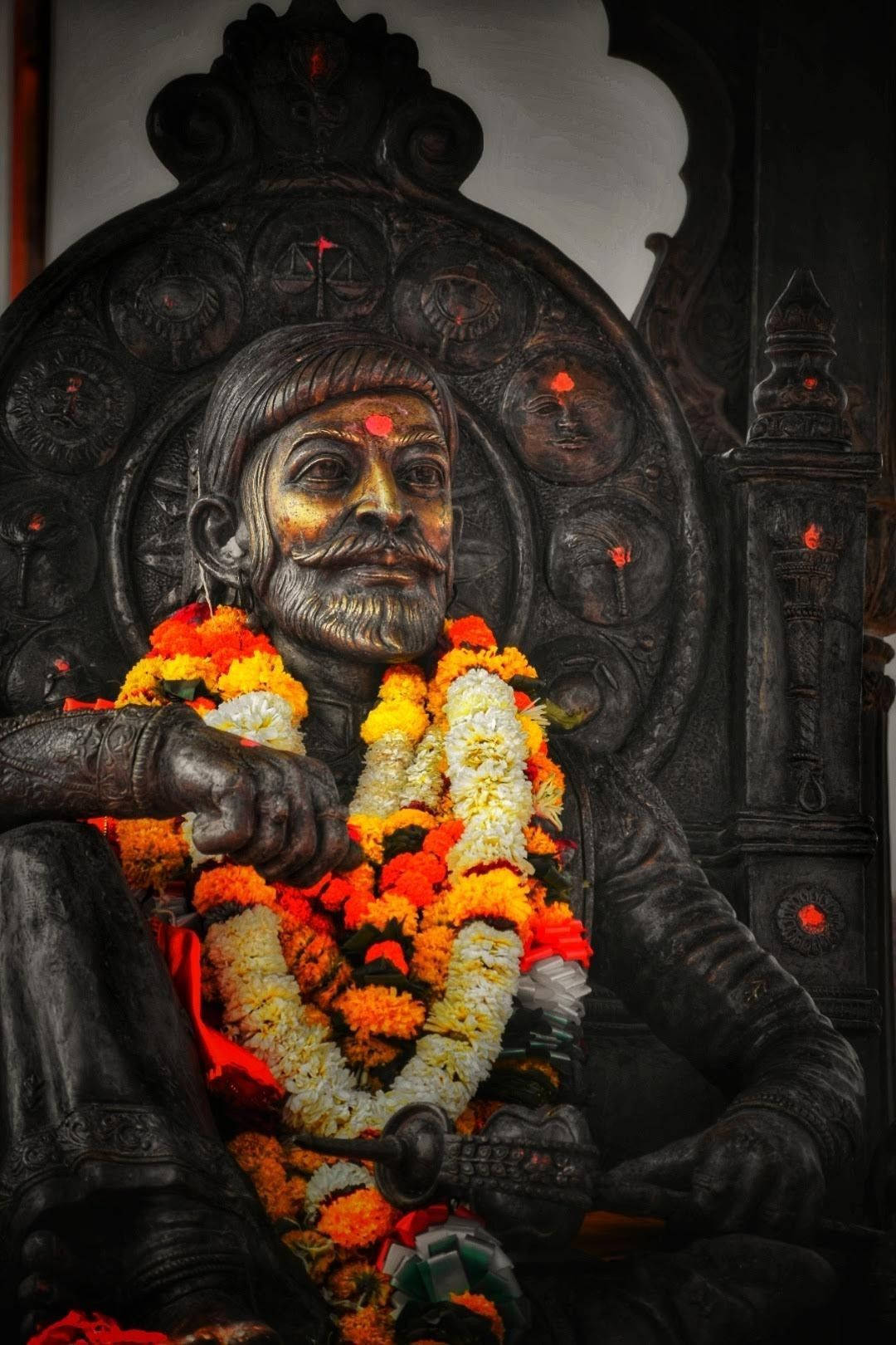 Chhatrapati Shivaji Maharaj 桌布 壁紙 — 一尊深色青銅鬍鬚男子雕像，飾以鮮豔的橙色和白色花環，莊嚴地坐在華麗的寶座上。