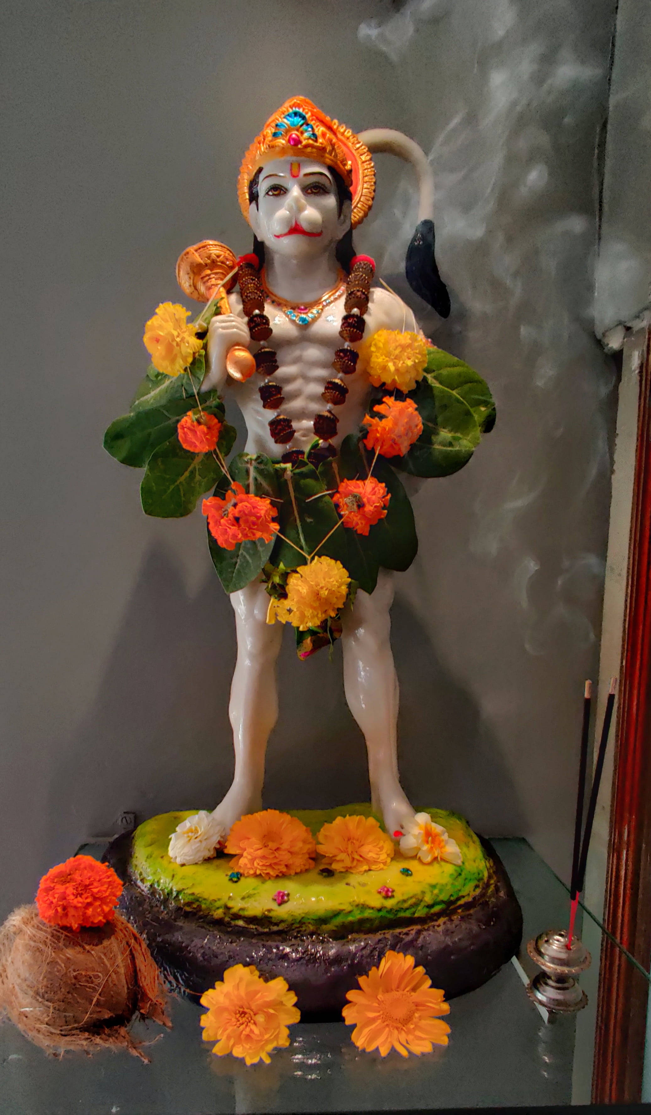 lord hanuman 桌布 壁紙 — 一尊白色哈努曼雕像，飾以橙色萬壽菊和綠葉，立於裝飾底座上，附近有香煙裊裊升起。