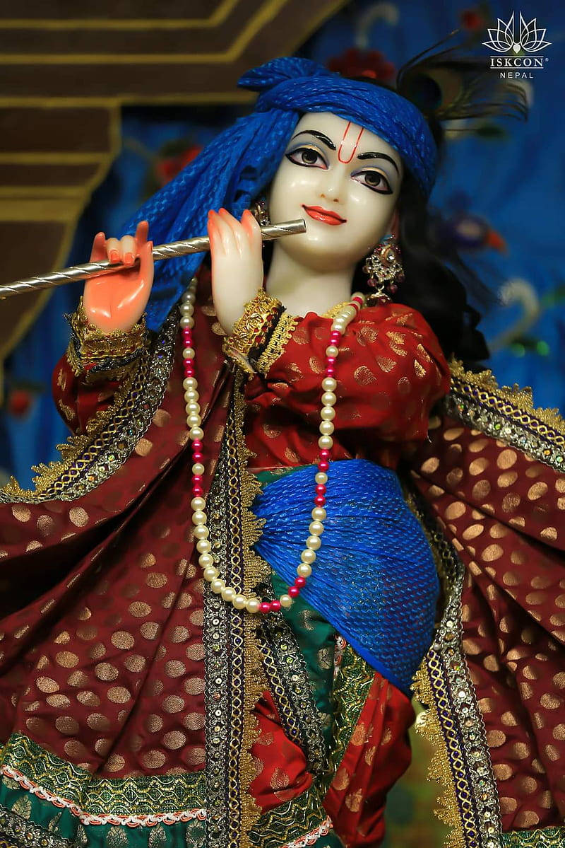 krishna god 桌布 壁紙 — 一尊神聖的克里希納雕像，身著鮮豔的紅色和藍色服飾，在寺廟背景下以寧靜的表情吹奏長笛。