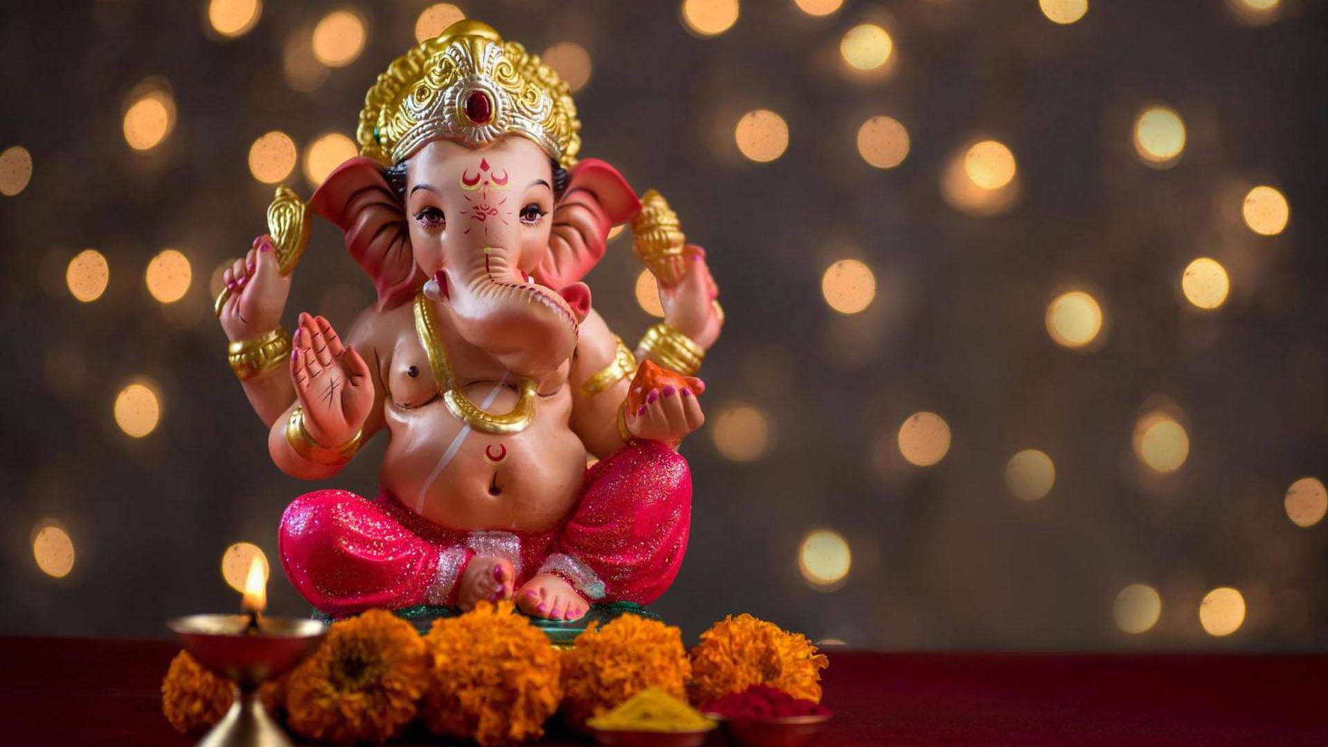 Hindu God 桌布 壁紙 — 一尊色彩繽紛的象神雕像，飾以金色和粉紅色，伴隨著萬壽菊和點燃的油燈，在溫暖的散景背景前靜靜地坐著。