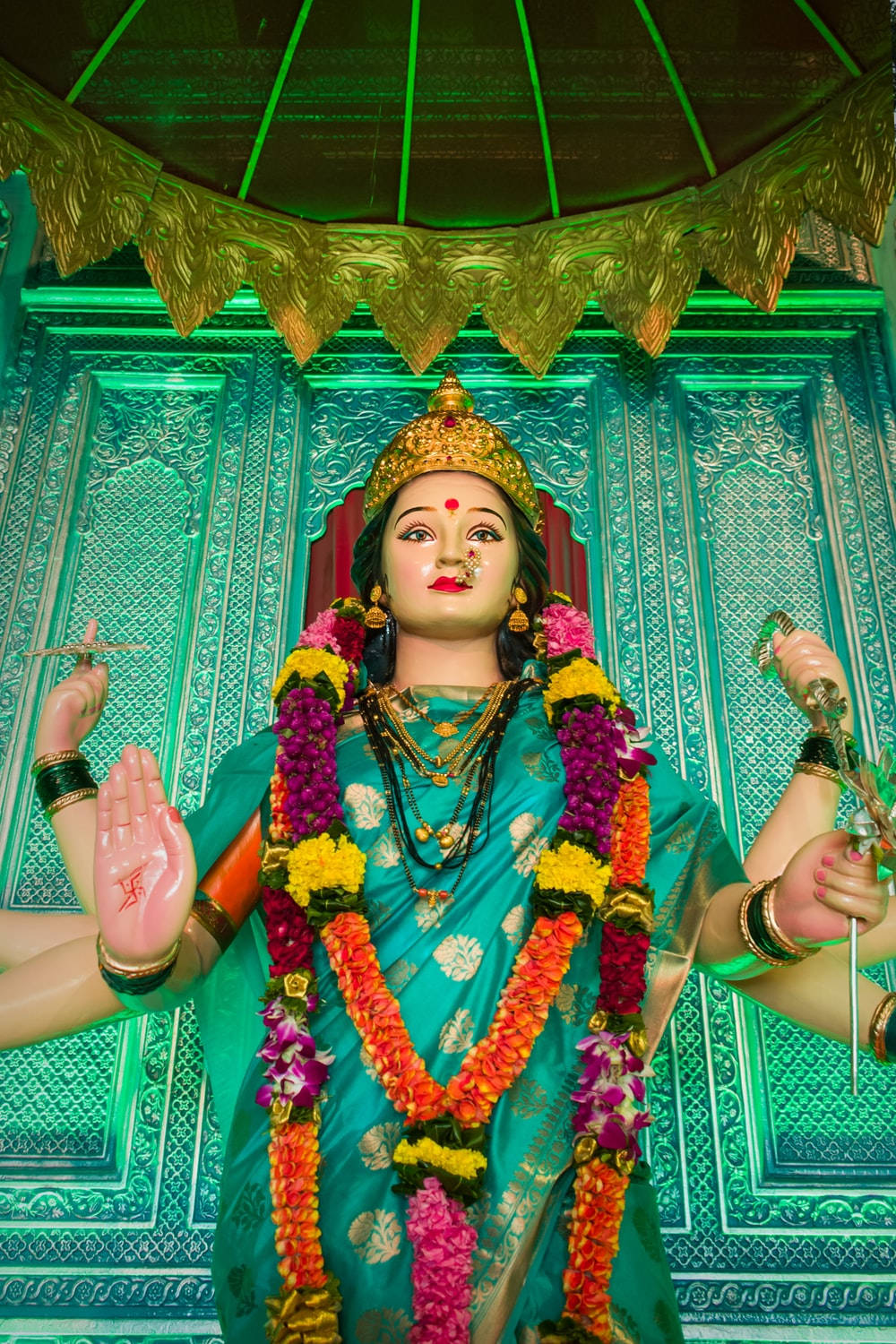 Vaishno Devi 桌布 壁紙 — 一尊色彩鮮豔的印度教女神雕像，飾有彩色花環和金飾，矗立在綠色燈光裝飾的華蓋下。