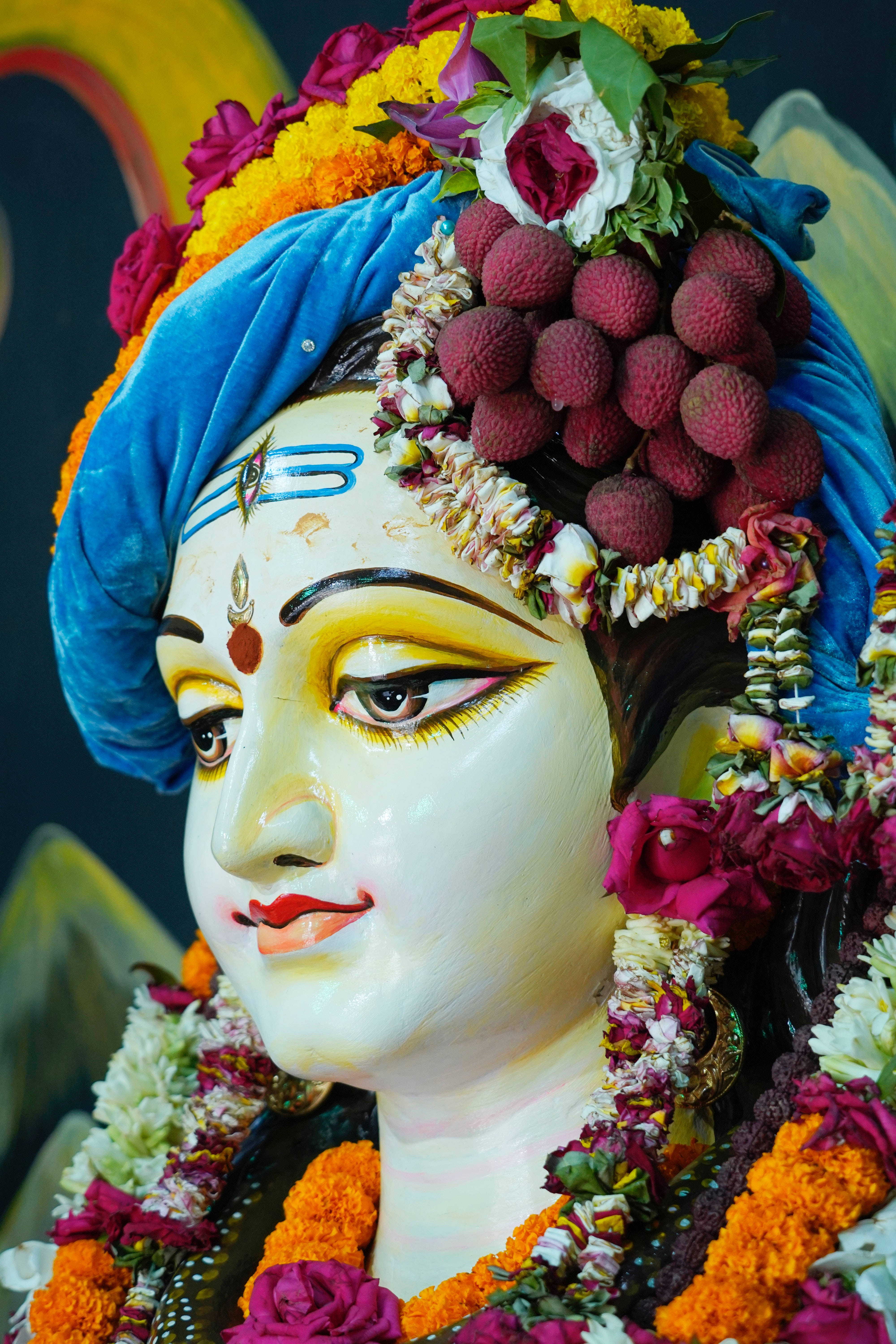 krishna iPhone 桌布 壁紙 — 一尊色彩鮮豔的神像，飾有彩色花朵、荔枝和藍色頭巾，平靜地凝視前方。