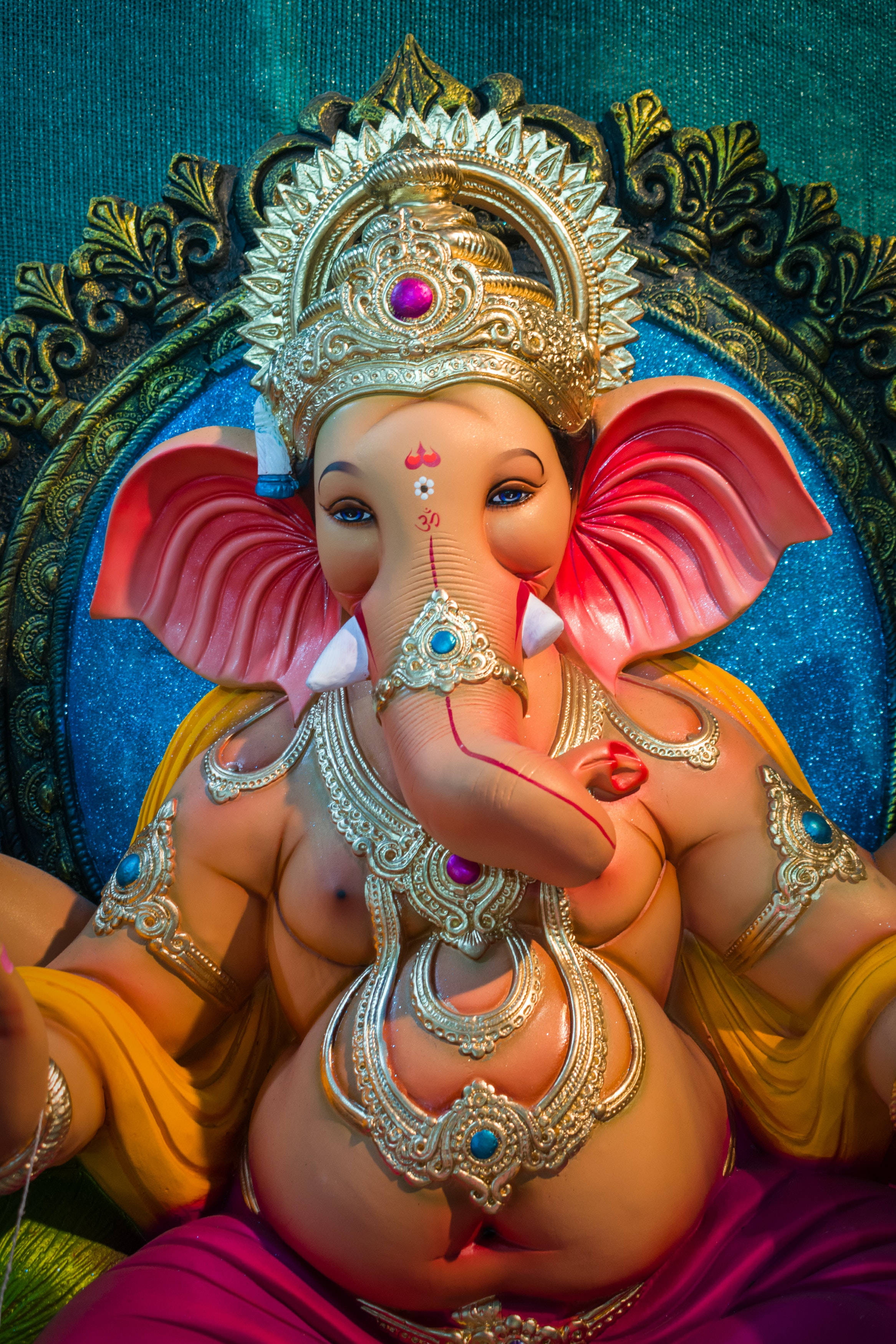 Ganesh Chaturthi 桌布 壁紙 — 一尊色彩鮮豔的象頭神像，飾有黃金珠寶和鑲寶石的王冠，莊嚴地坐在藍綠色和金色的華麗背景前。