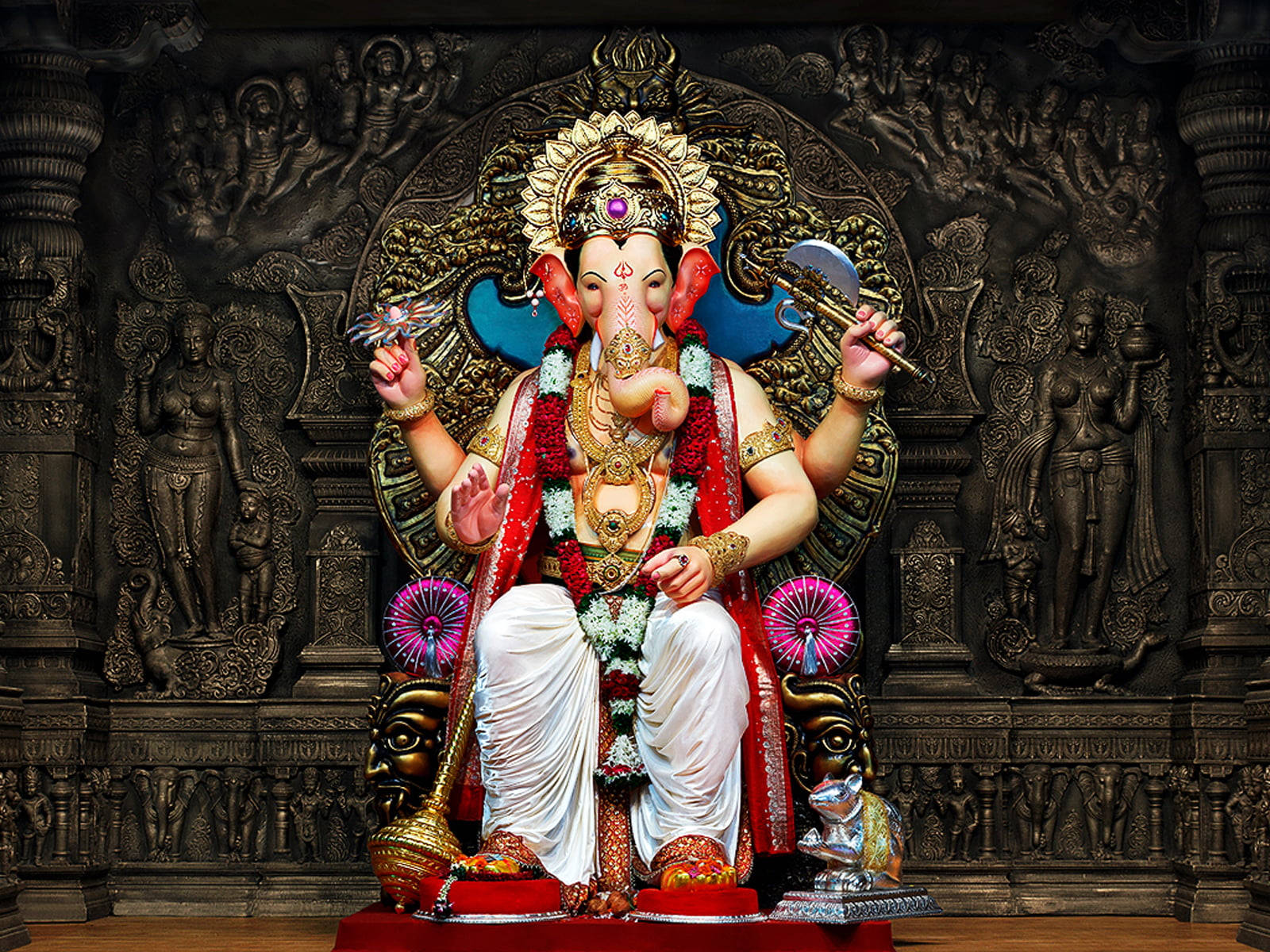 Ganesh 桌布 壁紙 — 一尊莊嚴的金色象神像，飾以紅色花環和珠寶，威嚴地坐在華麗的寶座上，背景是深色雕刻的寺廟。
