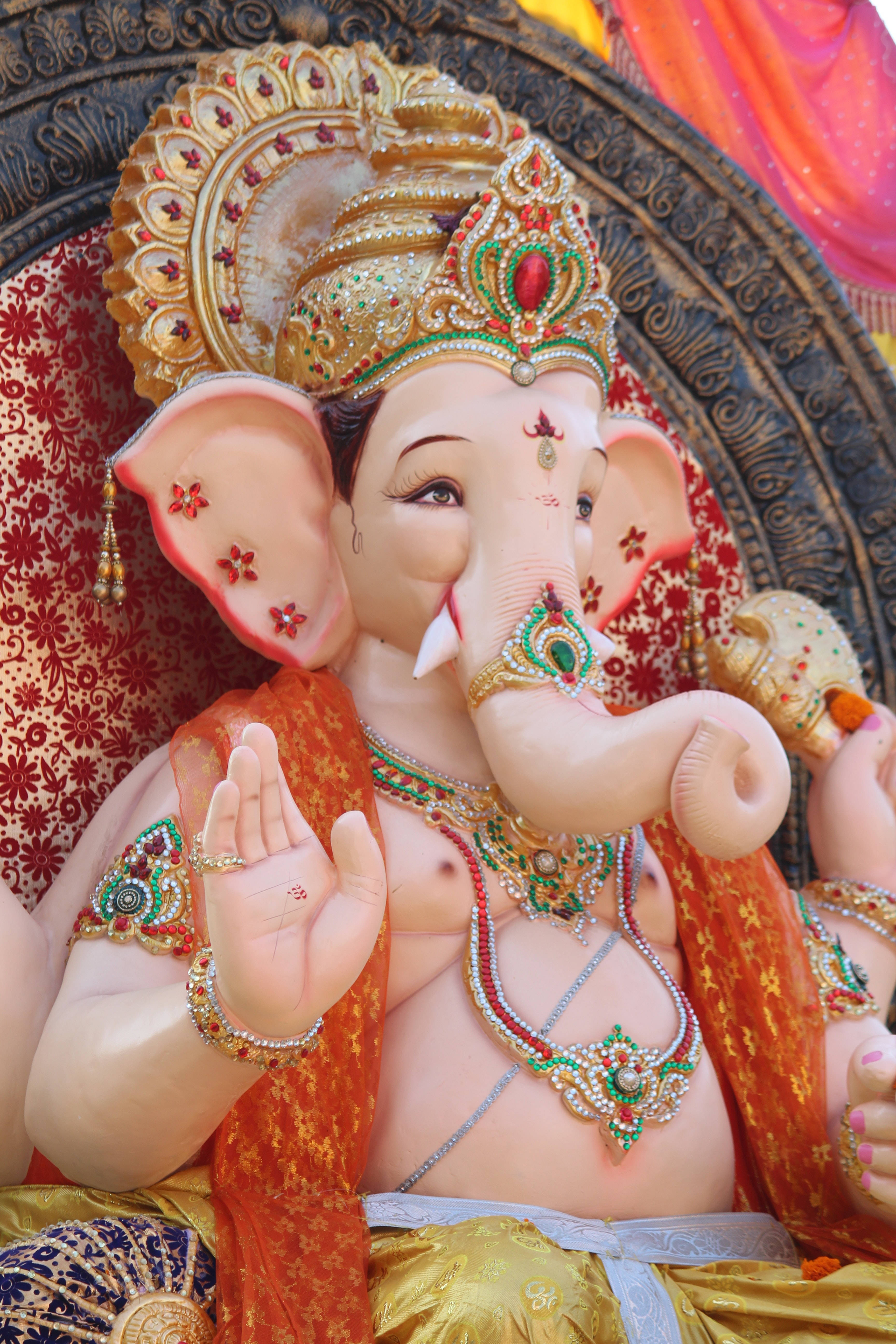 ganesh iPhone 桌布 壁紙 — 一尊裝飾華麗的象頭神像，頭戴金色皇冠，飾有紅綠寶石，披著橙色布料，莊嚴地坐在華麗圖案背景前。