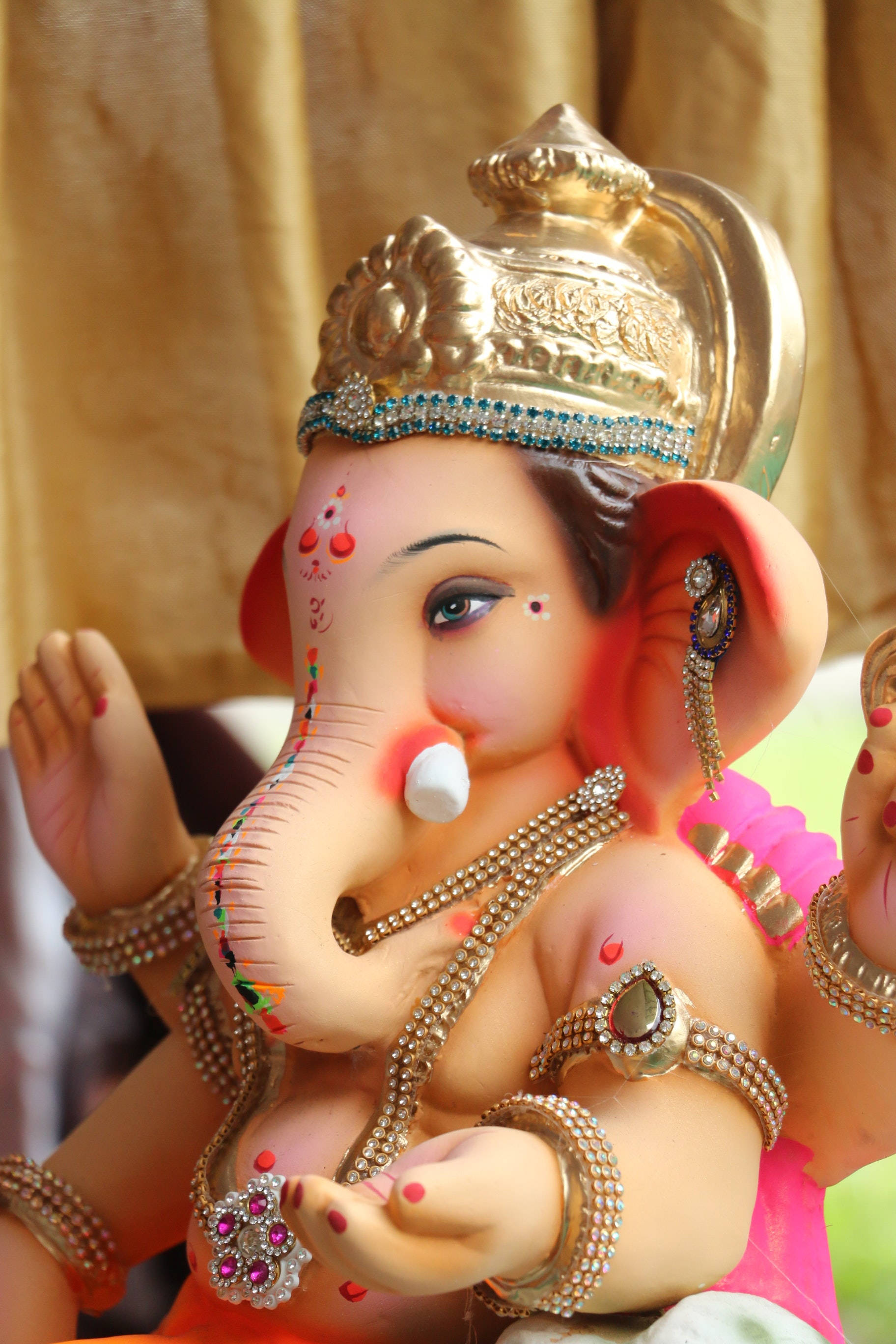 ganesh iPhone 桌布 壁紙 — 一尊裝飾華麗的象頭神雕像，飾有黃金首飾和彩色眉心貼，背景是金色窗簾。