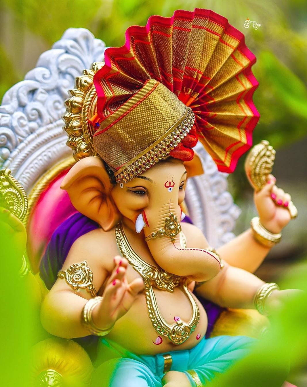 hindu iPhone 桌布 壁紙 — 一尊裝飾鮮豔的象頭神像，佩戴金色珠寶和紅色扇形皇冠，寧靜地坐在茂密的綠葉中。