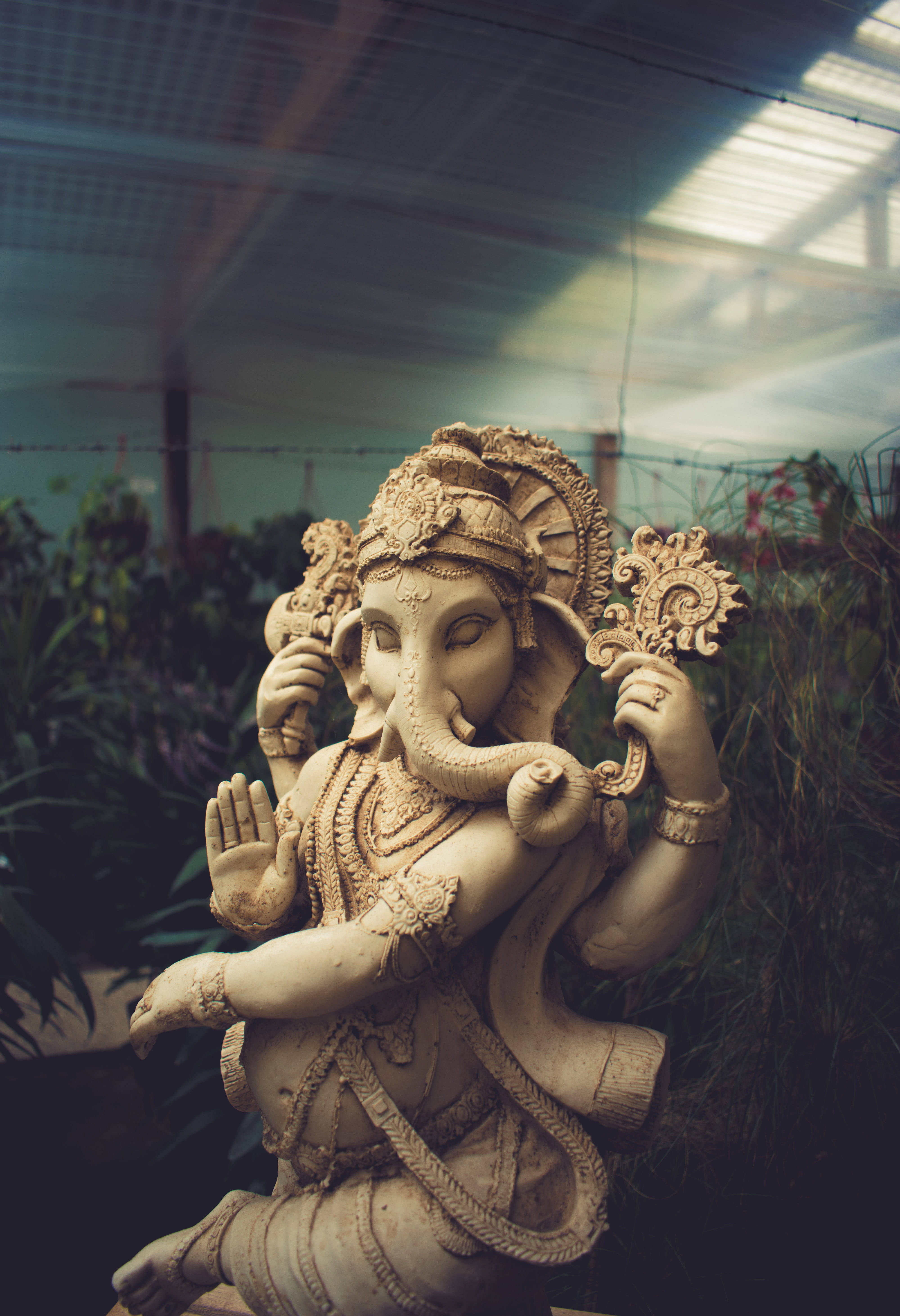 ganesh iPhone 桌布 壁紙 — 一尊詳細的象頭神伽內什石像，擁有象頭和華麗珠寶，在溫室屋頂下寧靜地坐於茂密的綠色植物之中。