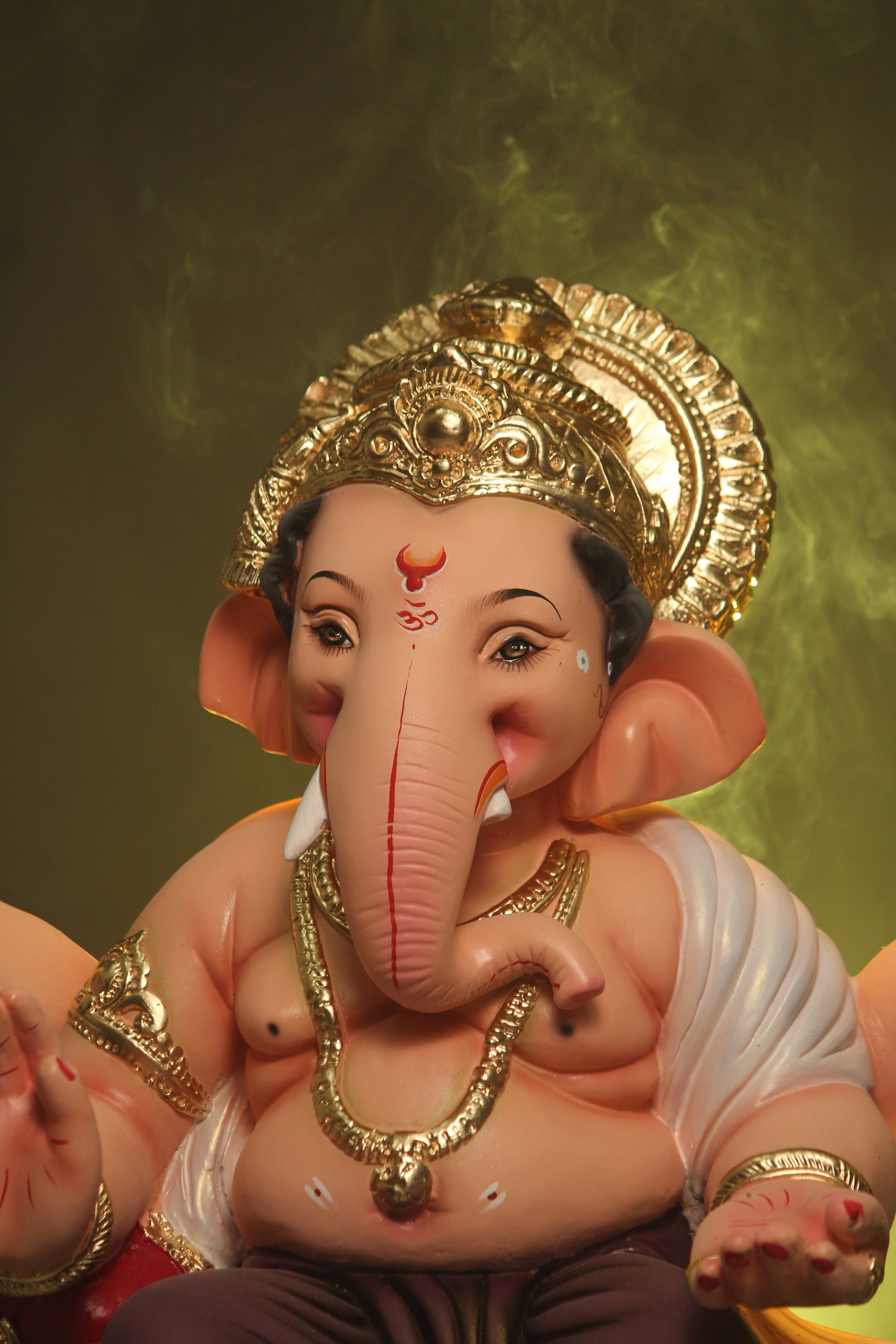 ganesh iPhone 桌布 壁紙 — 一尊詳細的象頭神雕像，飾有黃金珠寶和王冠，坐在煙霧繚繞的綠色背景前。