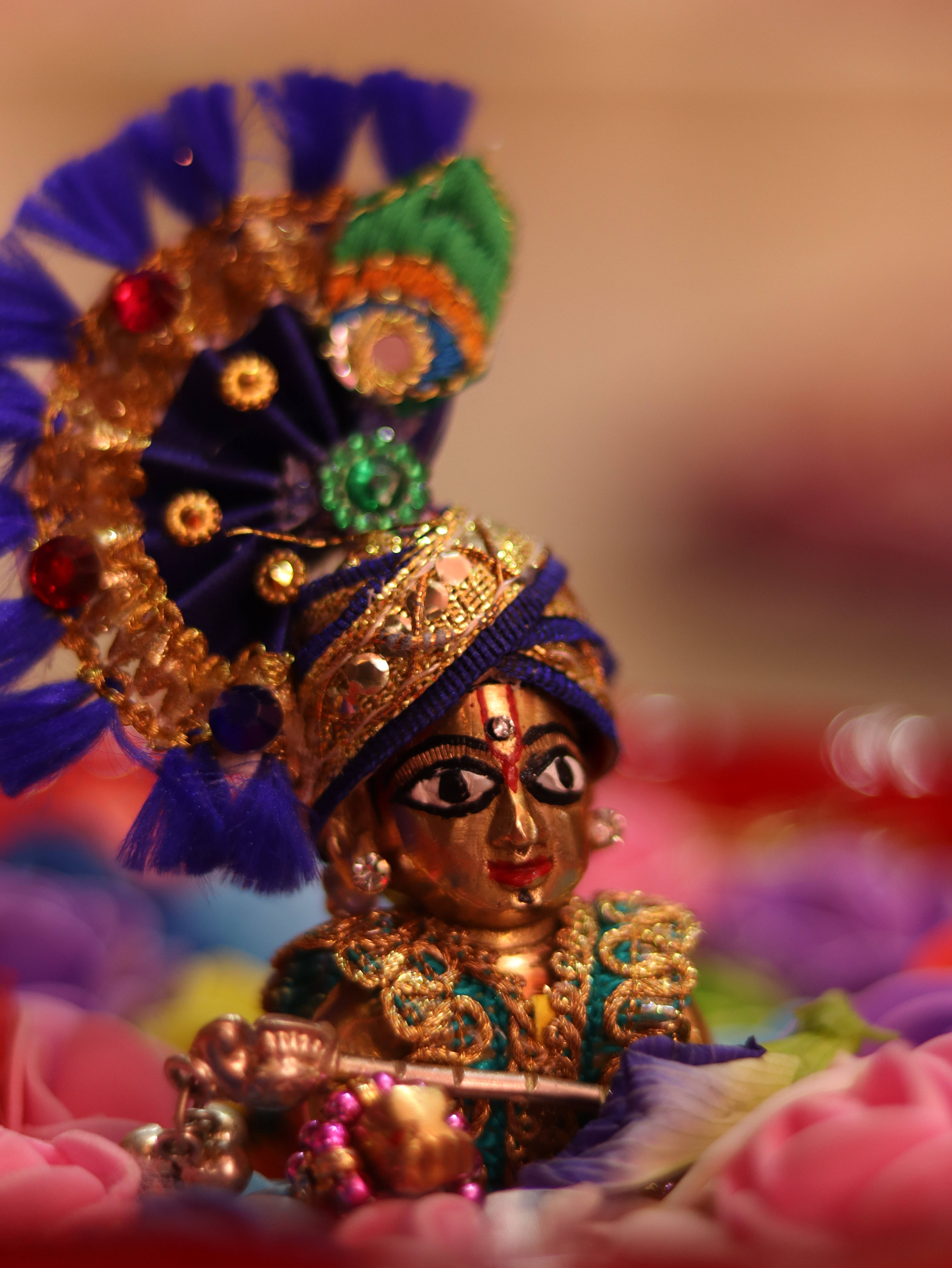 krishna iPhone 桌布 壁紙 — 一尊金色神像，飾有鑲嵌寶石的藍色頭飾，手持長笛，安放在色彩繽紛的花朵之中。