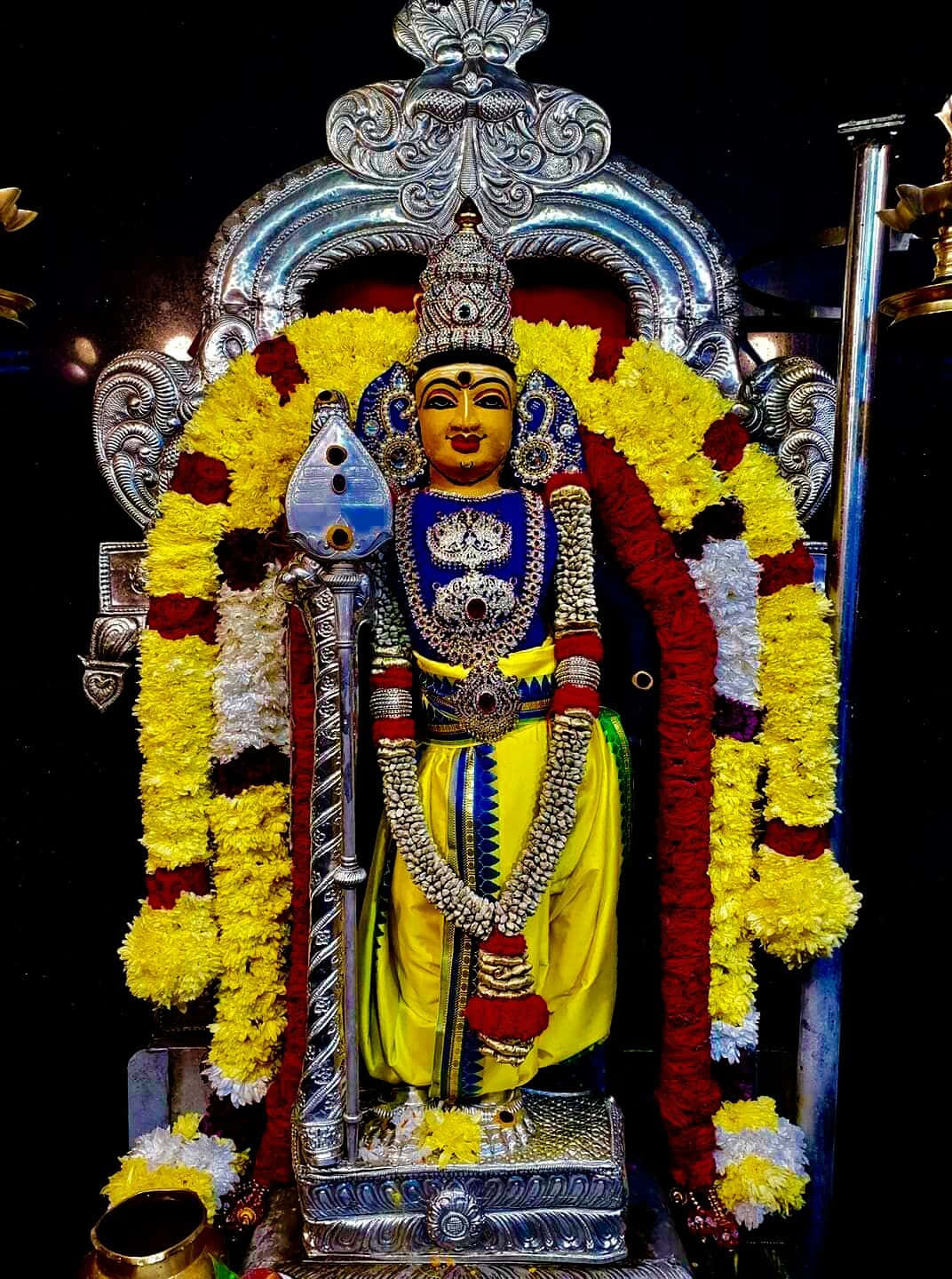 Murugan 桌布 壁紙 — 一尊閃耀的印度教神像，飾以精緻的銀飾和鮮豔的黃、紅、白色花環，矗立在裝飾拱門下。