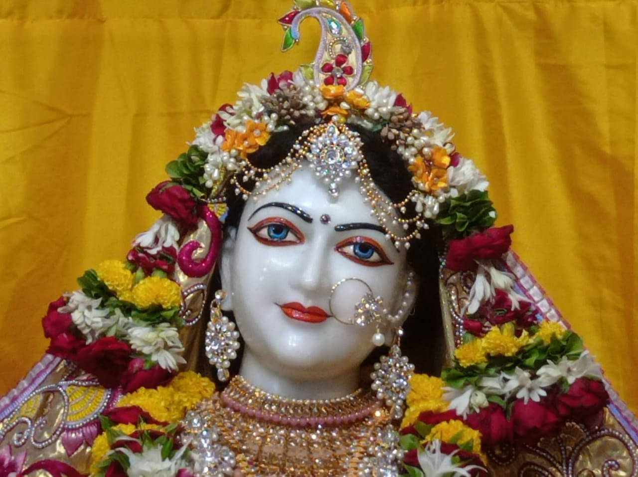 Hindu God 桌布 壁紙 — 一尊閃耀的白色神像，飾以色彩繽紛的花朵、珠寶和孔雀羽毛冠冕，襯以金色背景。