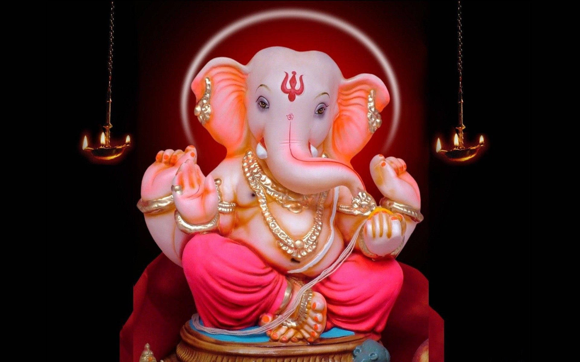 Hindu God 桌布 壁紙 — 一尊閃耀的粉紅色象頭神雕像，飾有黃金珠寶，安詳地坐在紅色基座上，兩旁是發光的油燈，背景漆黑。