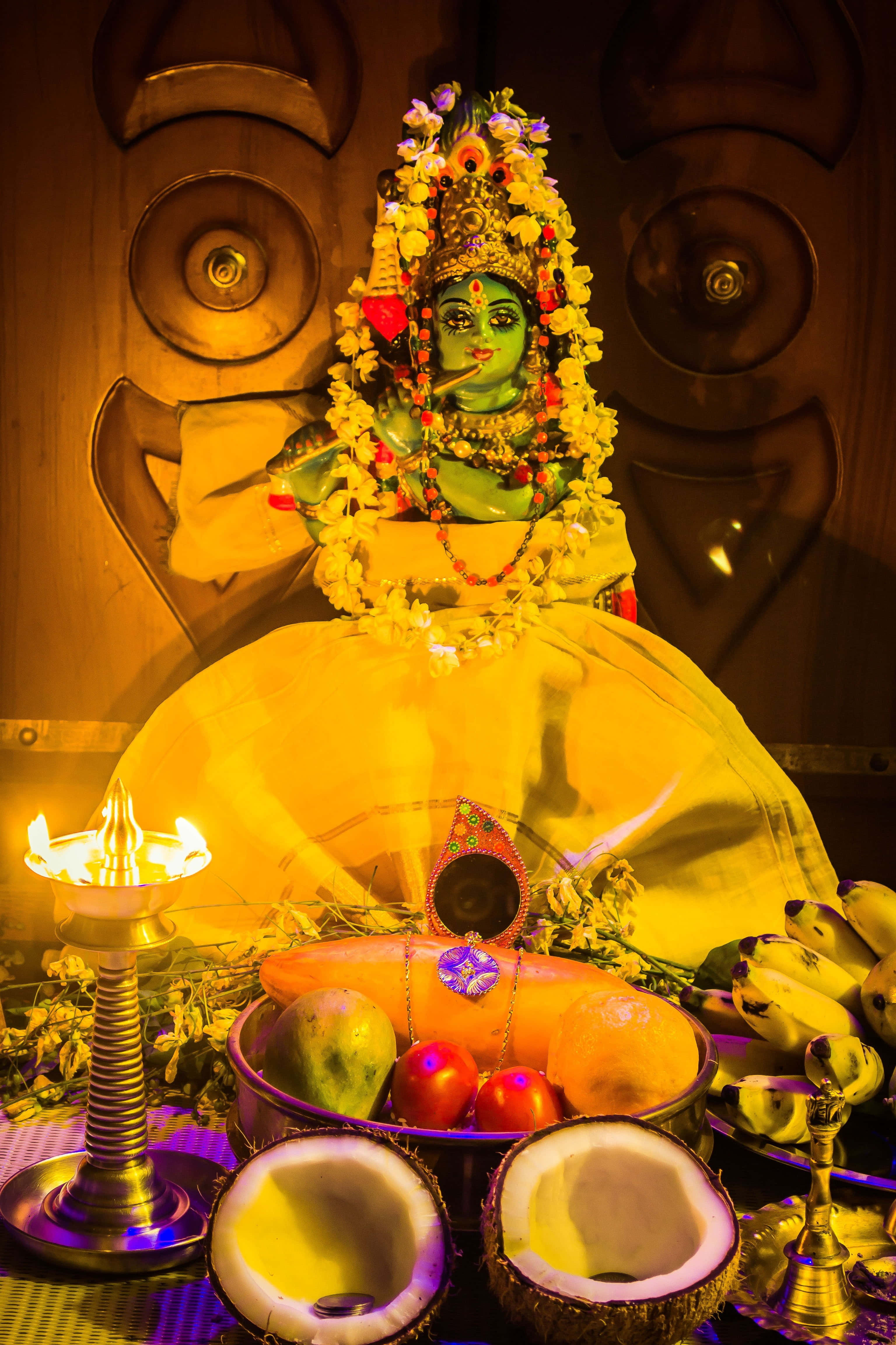 Vishu 桌布 壁紙 — 一尊閃耀的綠色神像，飾有鮮花和珠寶，坐在黃布上，周圍擺放著水果、椰子和點燃的油燈等供品。