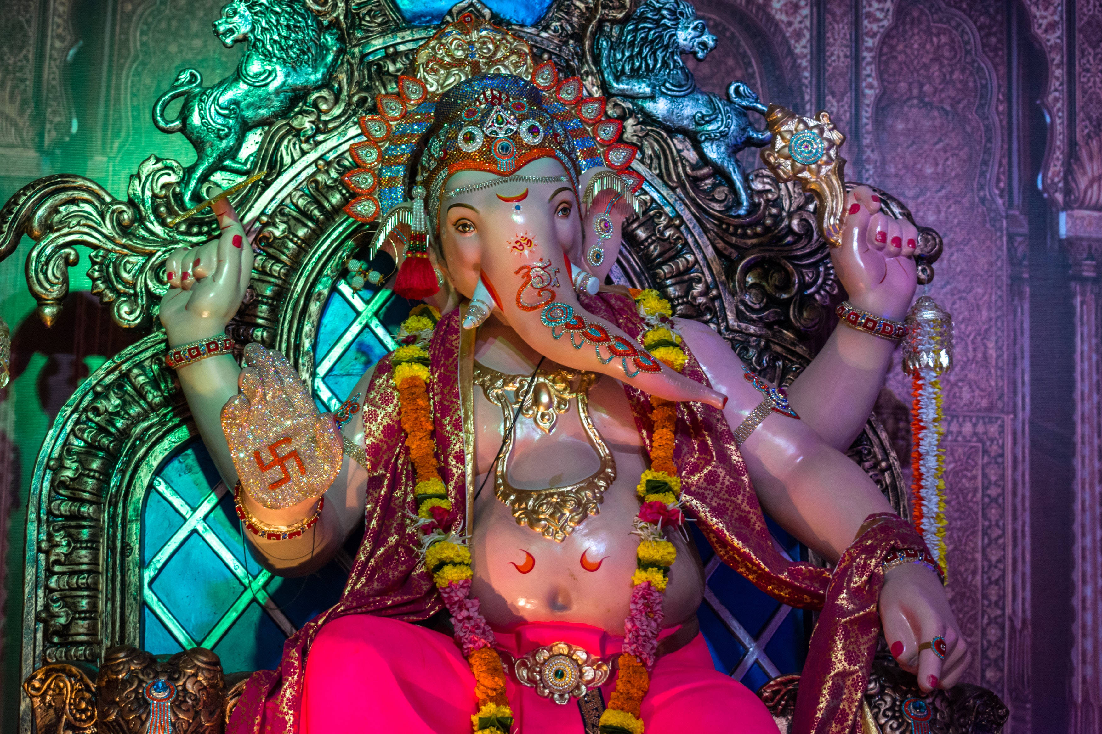 Ganesh 桌布 壁紙 — 一尊閃耀的象神雕像，飾有黃金珠寶、萬壽菊花環和紅色標記，在彩色燈光下莊嚴地坐在華麗的寶座上。