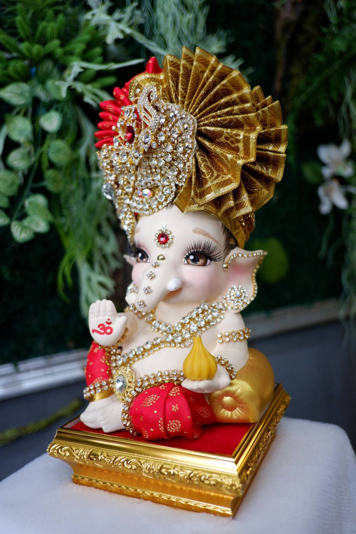 Baby Ganesh 桌布 壁紙 — 一尊閃閃發光、裝飾華麗的象頭神像，飾有金色和紅色寶石，優雅地坐在金色底座上，背景是茂密的綠色樹葉。