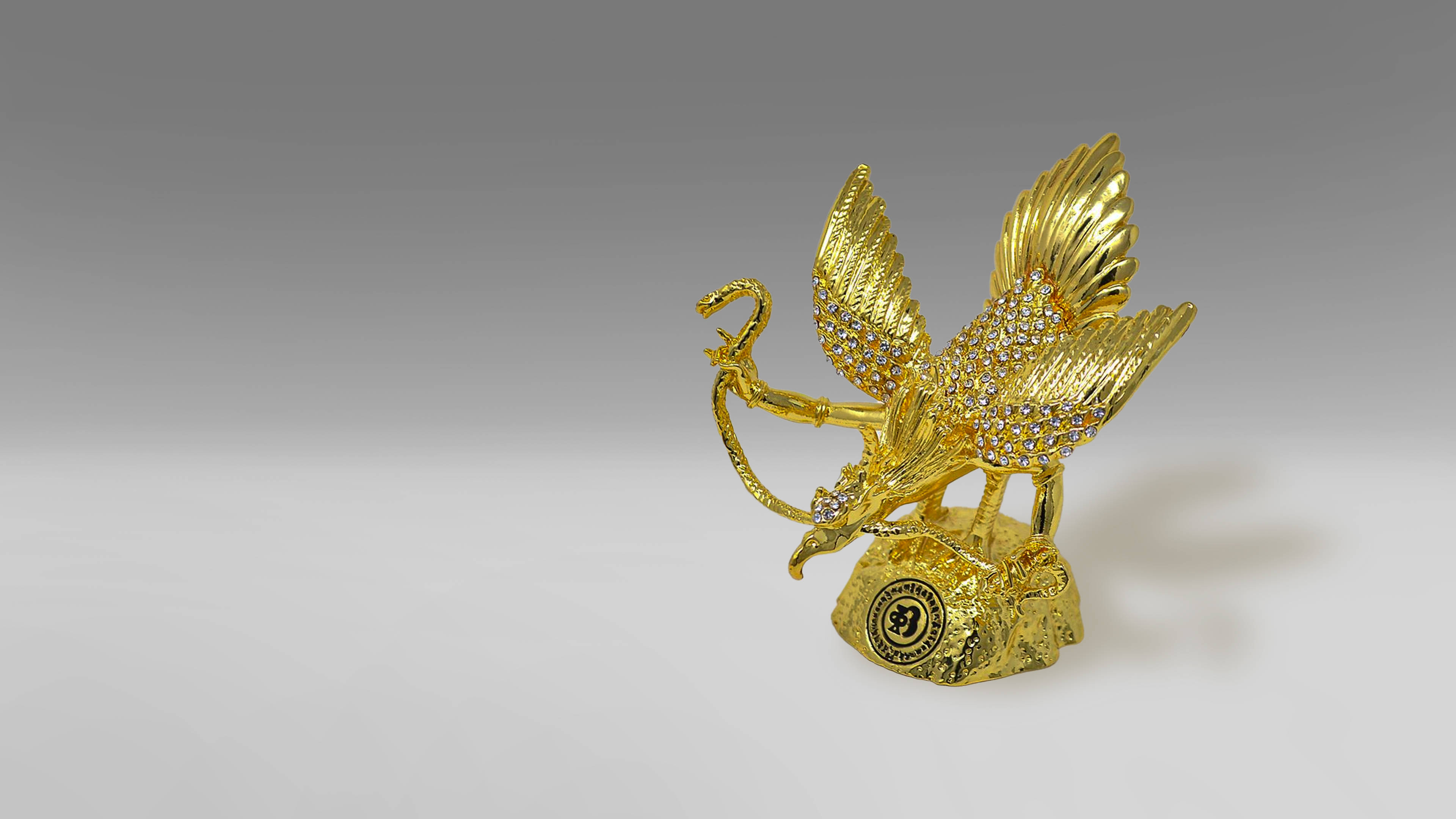 Garuda 桌布 壁紙 — 一尊閃閃發光的金色獅鷲雕像，鑲有寶石的翅膀，棲息在帶有紋理的基座上，背景是柔和的漸變色。