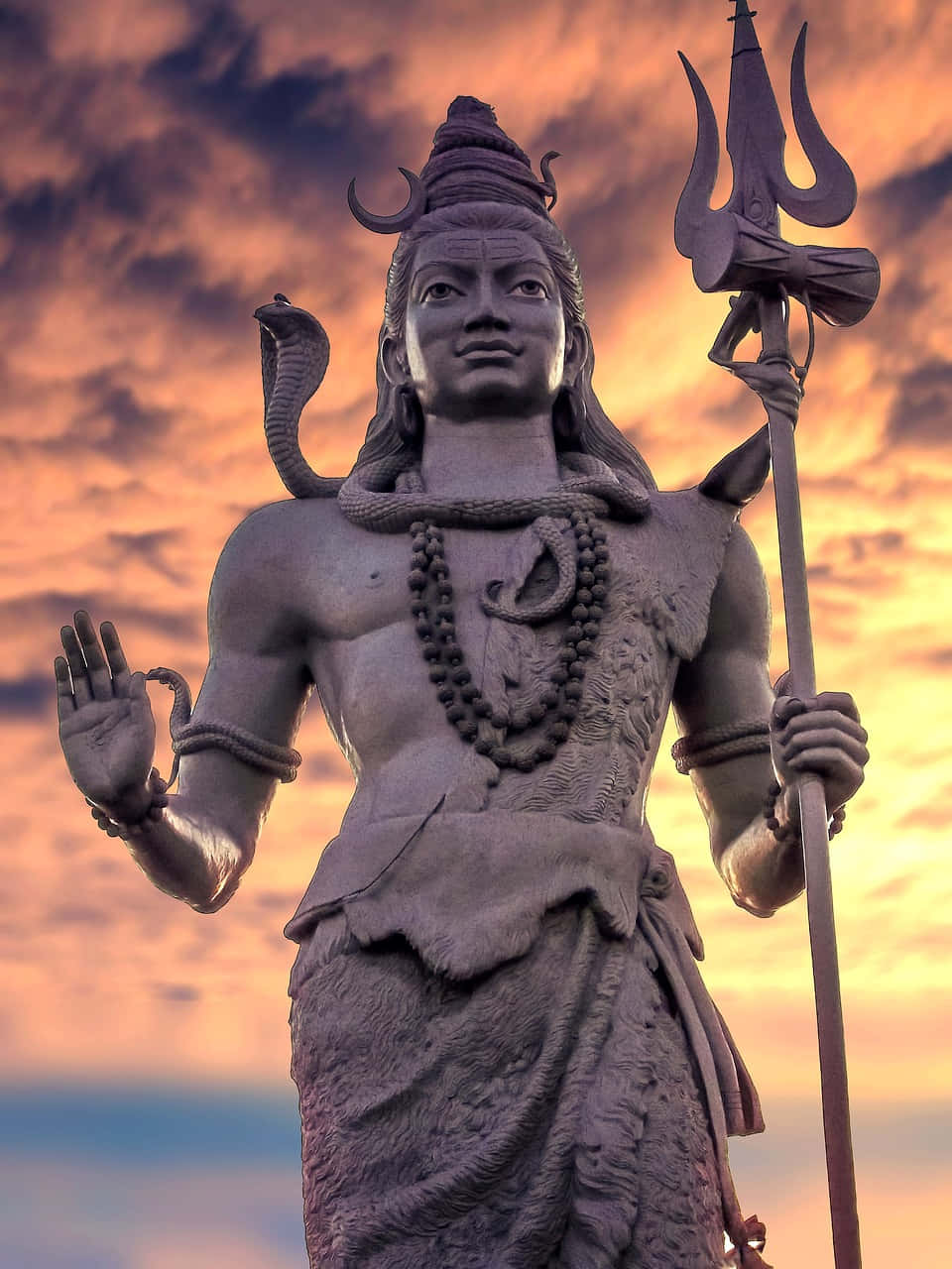 Mahadev 桌布 壁紙 — 一尊雄偉的濕婆神石像聳立在橙紫相間、雲彩閃耀的熾熱日落天空前。