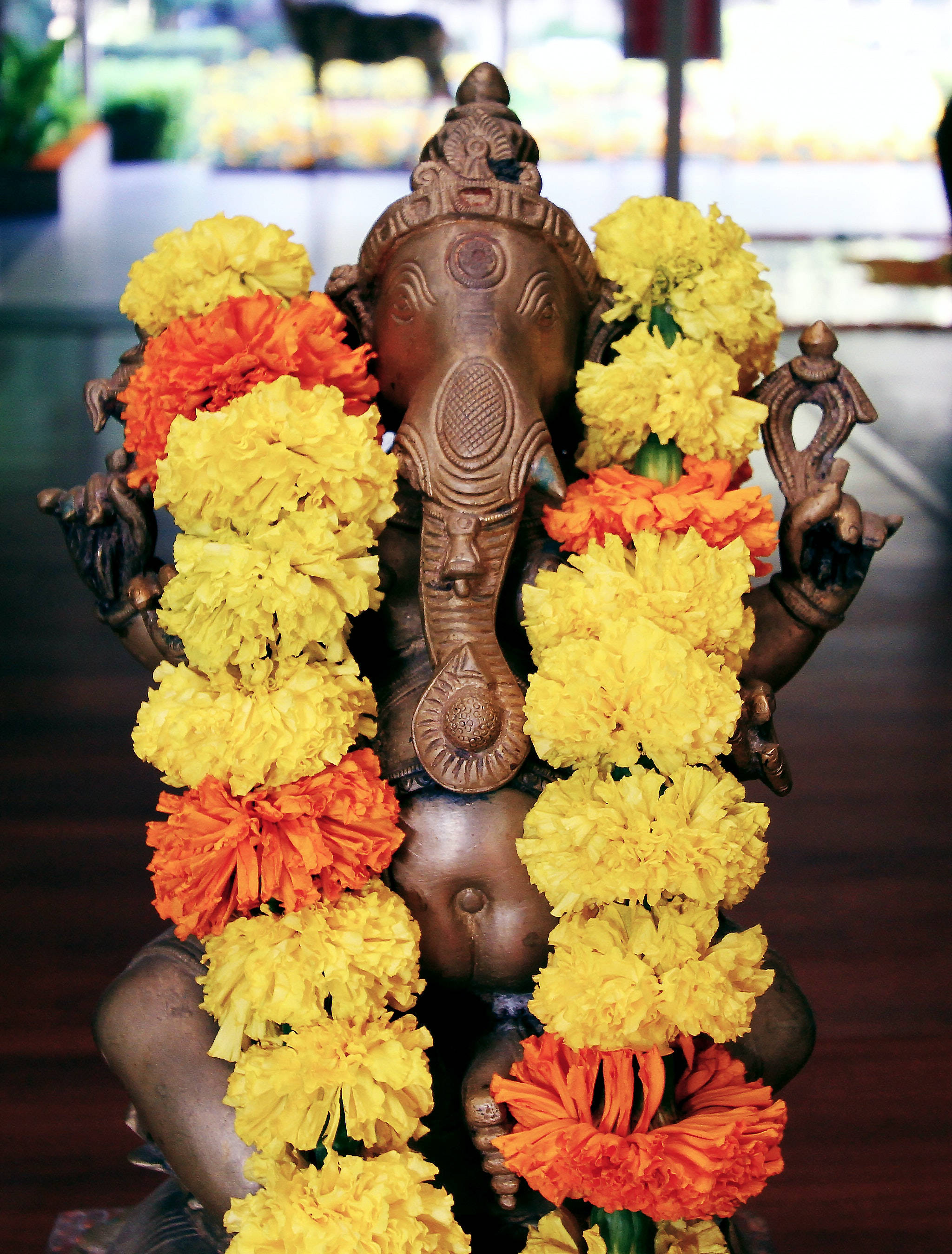 ganesh iPhone 桌布 壁紙 — 一尊青銅象神像，飾以鮮豔的黃色和橙色萬壽菊花環，置於室內，背景模糊。