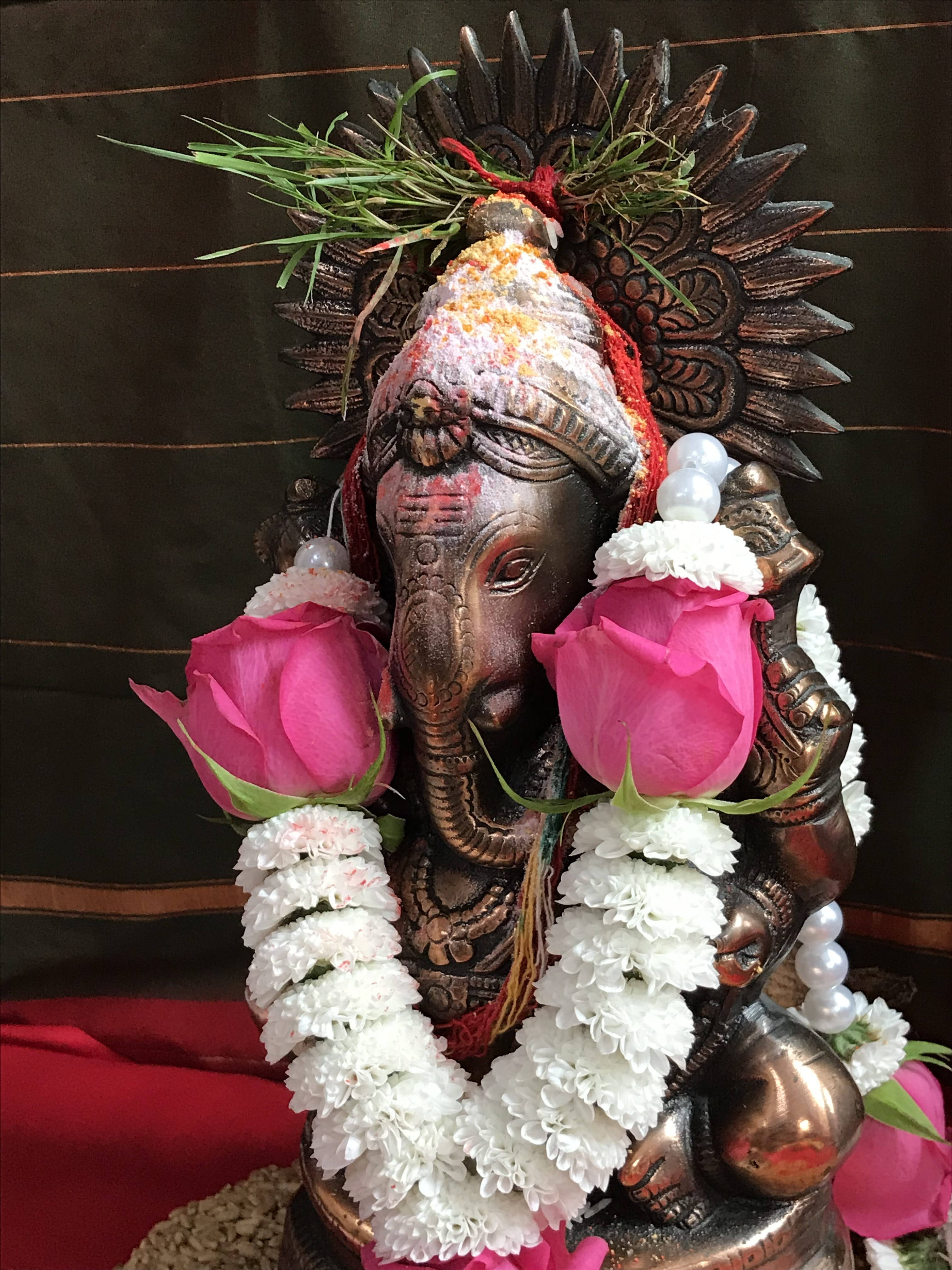 ganesh iPhone 桌布 壁紙 — 一尊青銅象神雕像，飾以粉紅玫瑰、白色花朵和珍珠，背景為深色垂簾。
