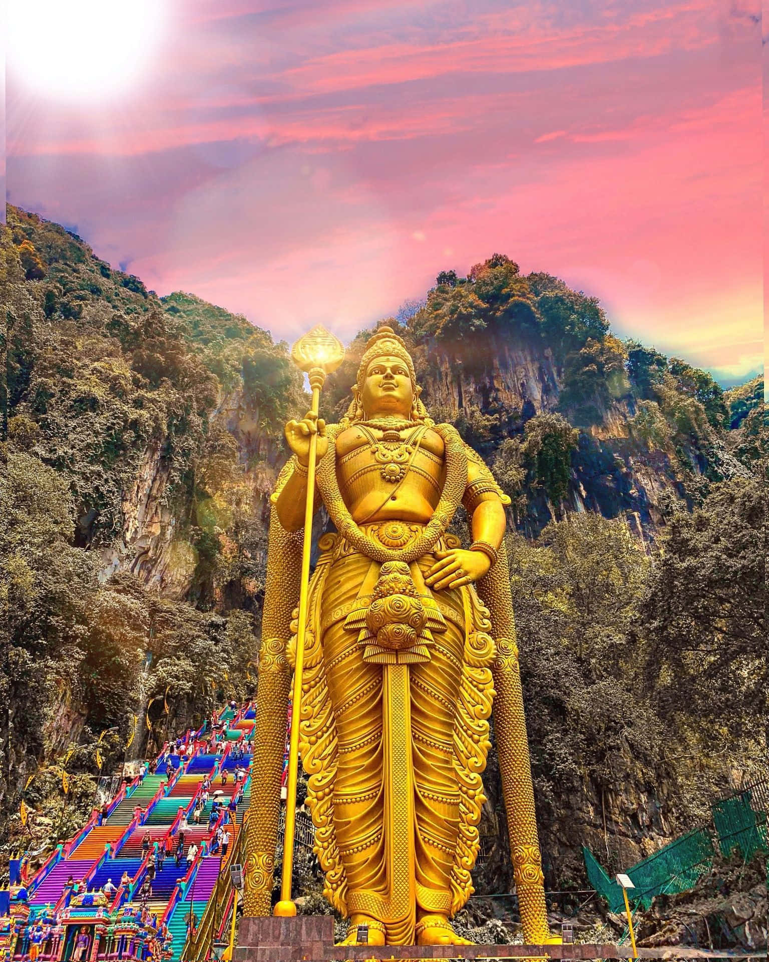 Murugan 桌布 壁紙 — 一尊高聳的穆魯干勳爵金色雕像，在充滿活力的粉紅色和橙色日落天空下，背後是鬱鬱蔥蔥的綠色山脈，莊嚴地矗立著。