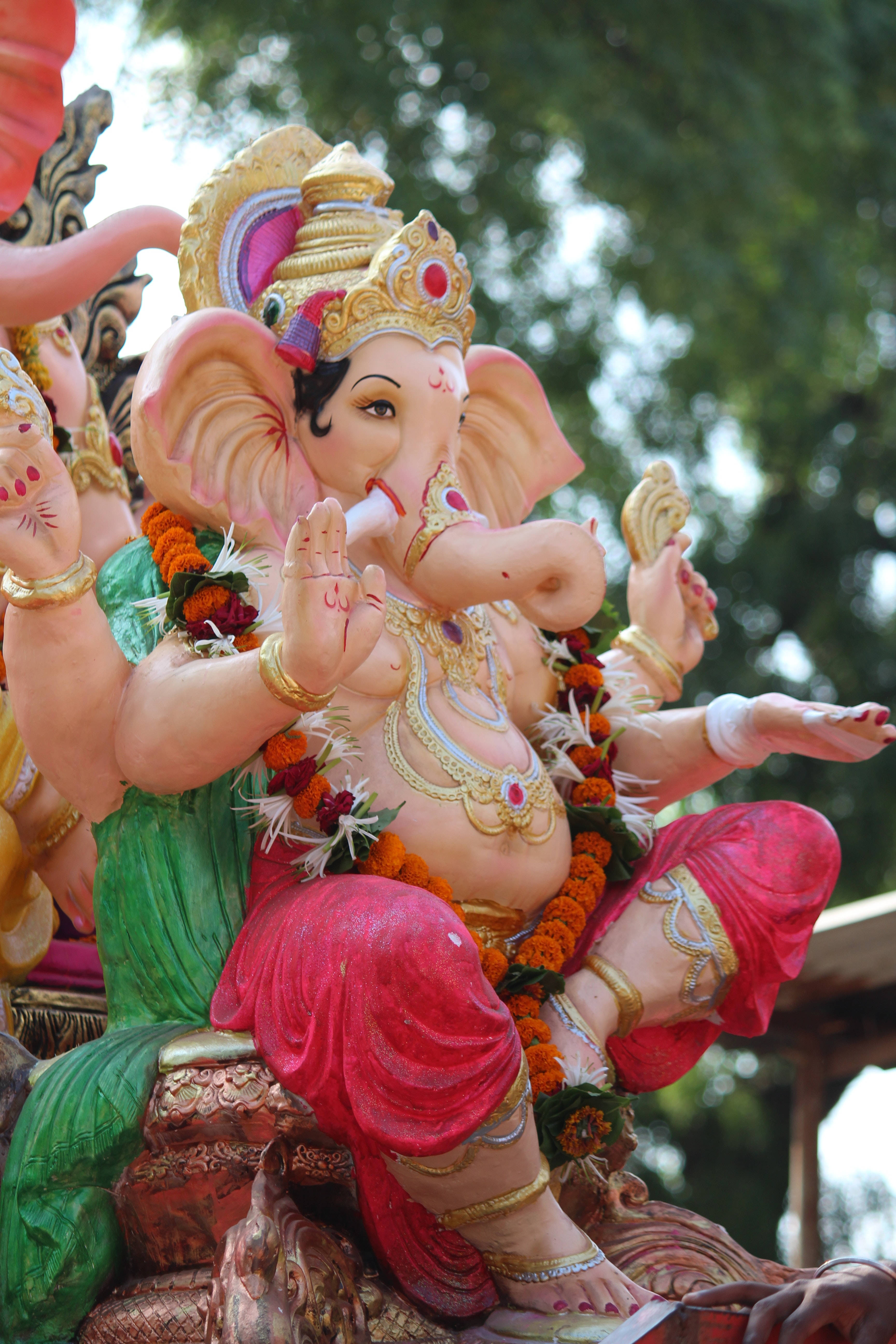 ganesh iPhone 桌布 壁紙 — 一尊鮮豔的粉紅色象頭神雕像，飾有黃金珠寶和金盞花花環，在斑駁的陽光下寧靜地坐在戶外。