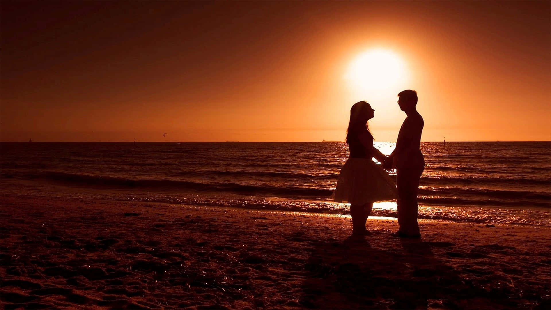 Beach Lovers 桌布 壁紙 — 一對剪影情侶手牽手站在沙灘上，夕陽在他們身後落下，在海面上投射出溫暖的橙色光芒。