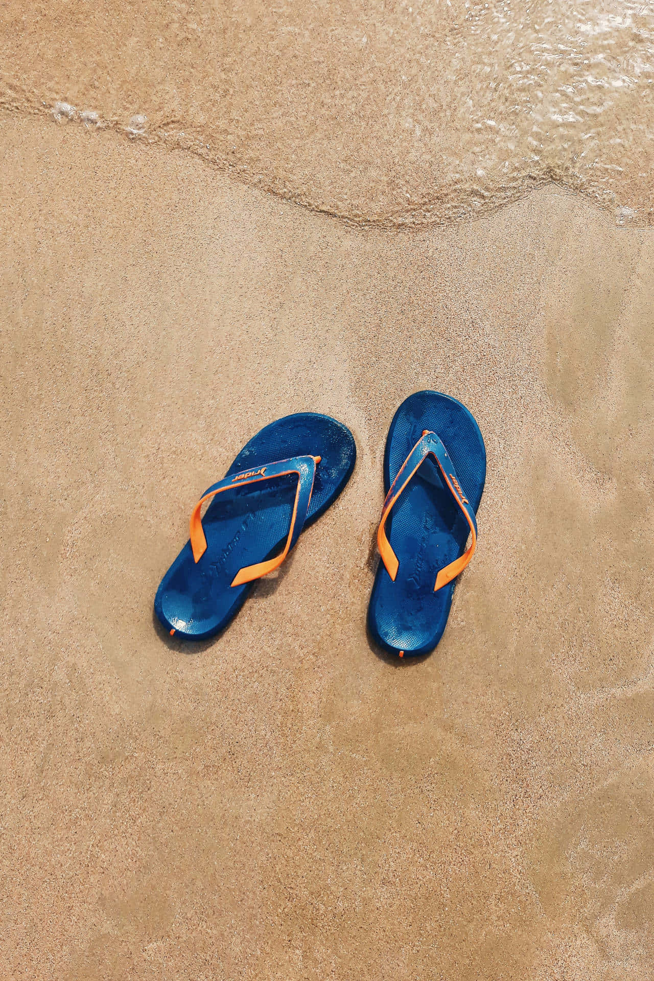 Beach Sandals 桌布 壁紙 — 一對帶有橙色帶子的藍色人字拖放在濕沙上，溫柔的海浪在附近拍打。