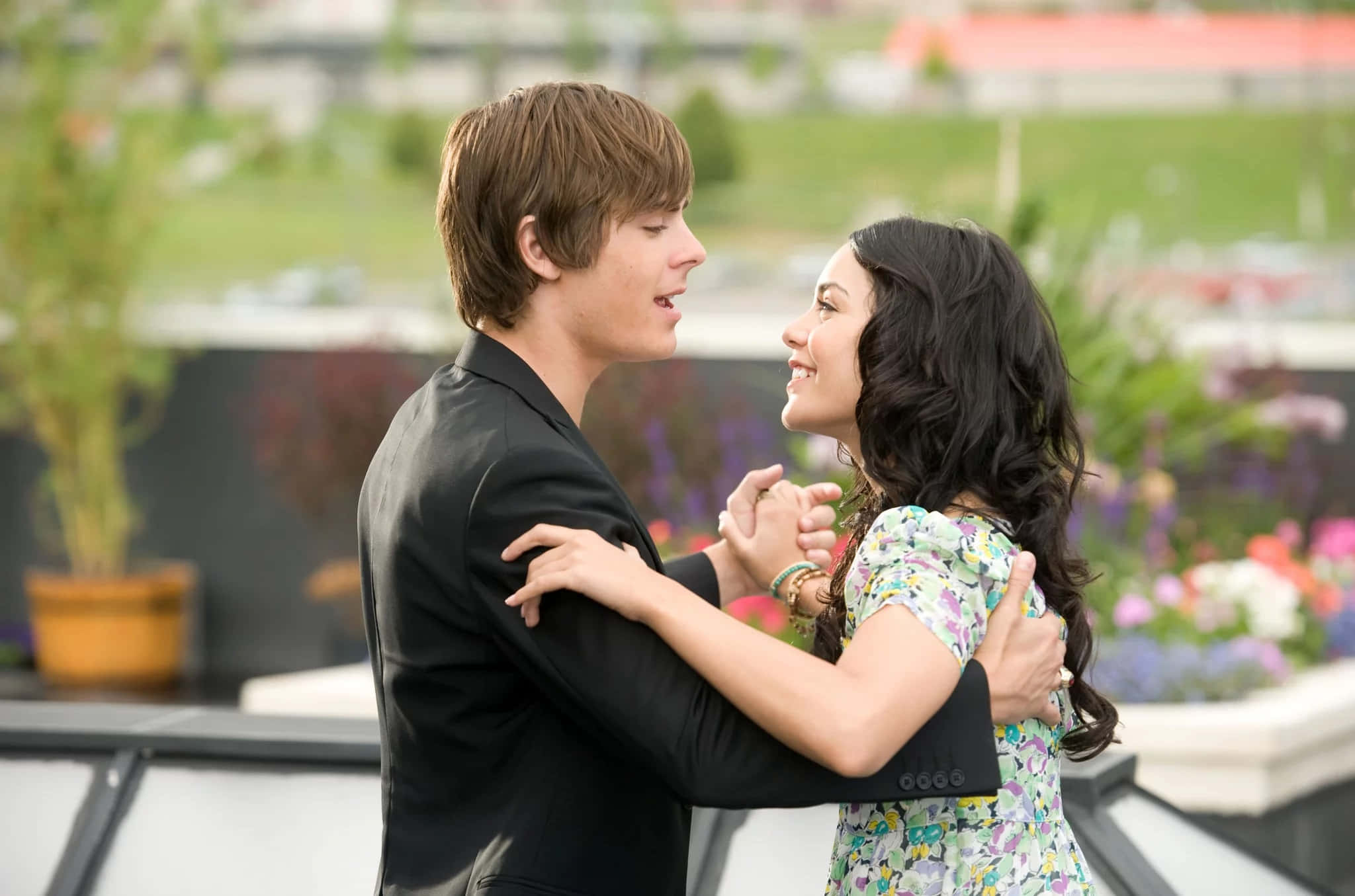 High School Musical 桌布 壁紙 — 一對年輕情侶在一個充滿鮮花和柔和日光的屋頂花園中親密共舞。