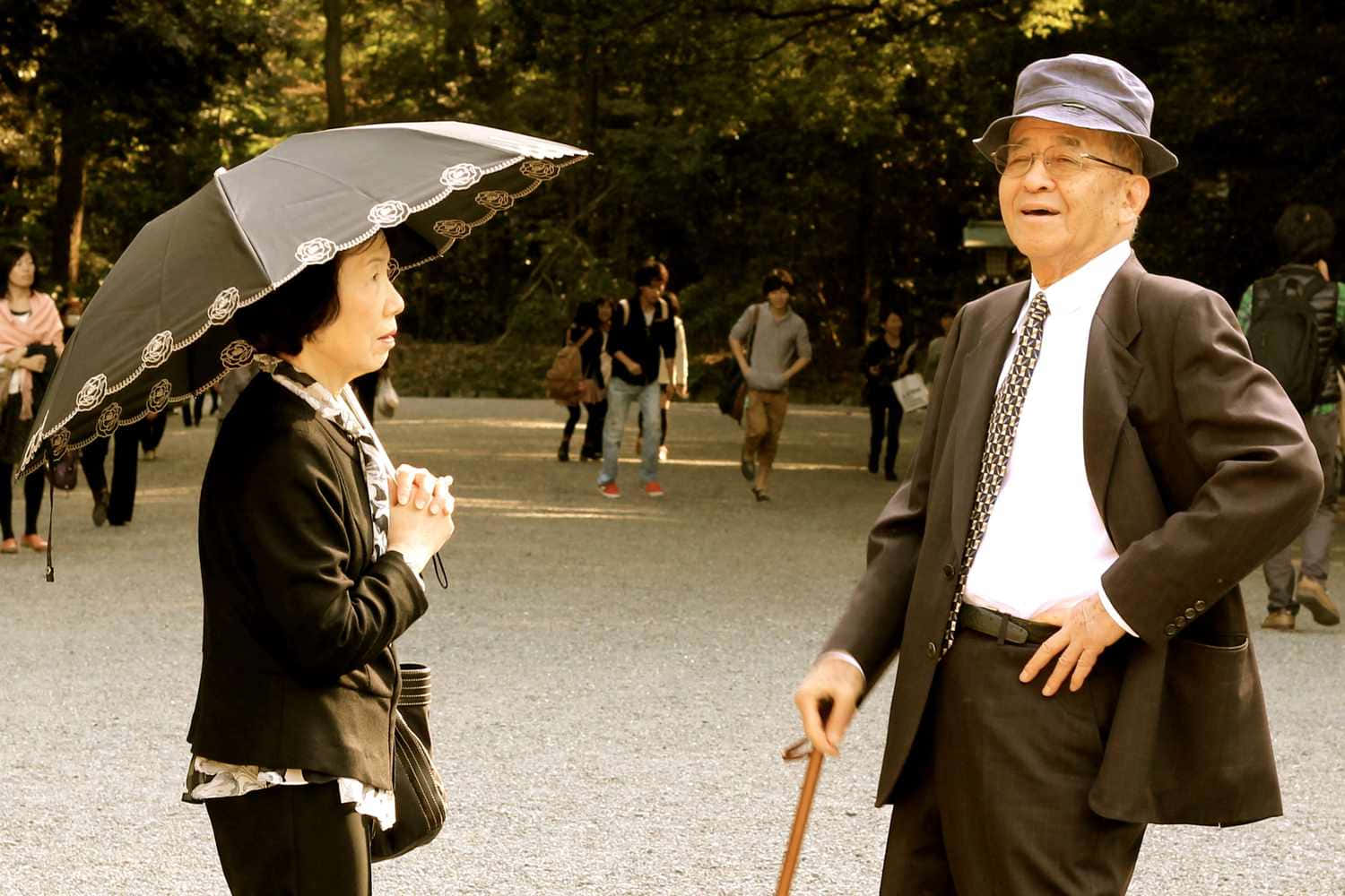Japanese 桌布 壁紙 — 一對年長夫婦在陽光明媚的公園裡共享寧靜時刻，女士手持華麗的雨傘，男士則倚靠著手杖。