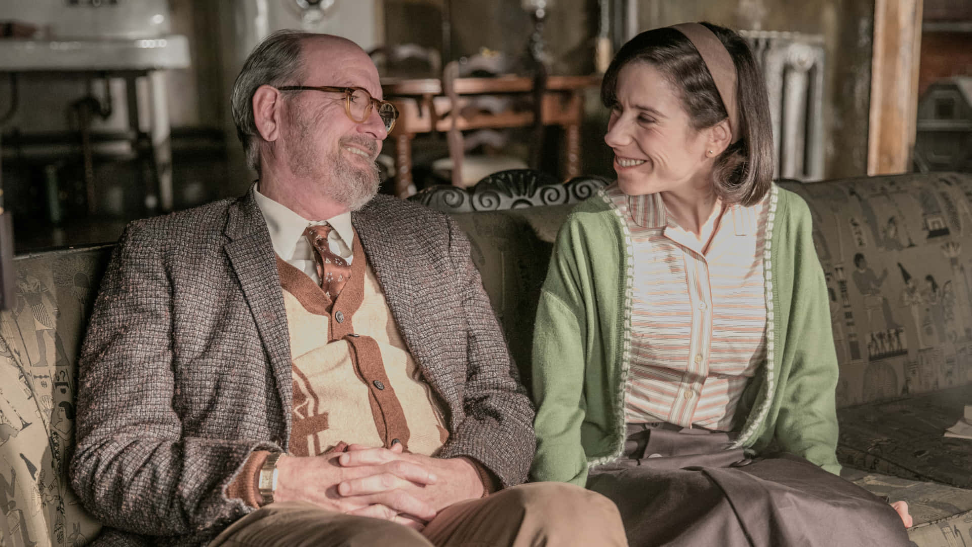 Richard Jenkins 桌布 壁紙 — 一對年長夫婦坐在復古風格房間的圖案沙發上，溫暖地互相微笑。