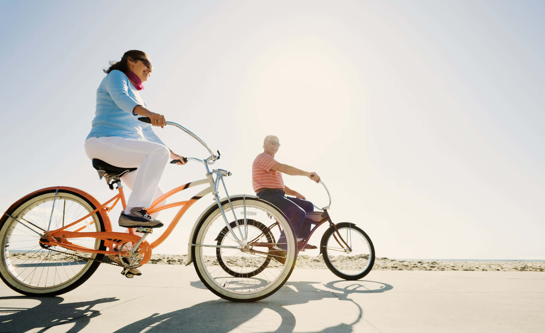 Beach Cruiser 桌布 壁紙 — 一對年長夫婦沿著陽光燦爛、天空清澈、影子長長的海灘騎著復古單車。