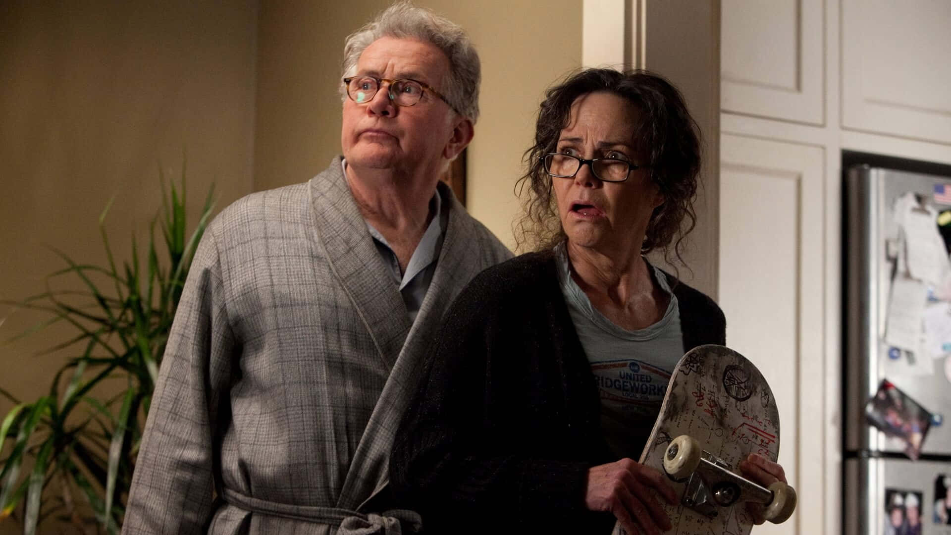 Aunt May 桌布 壁紙 — 一對年長夫婦穿著浴袍站在廚房裡，驚訝地向上看，女士拿著滑板。