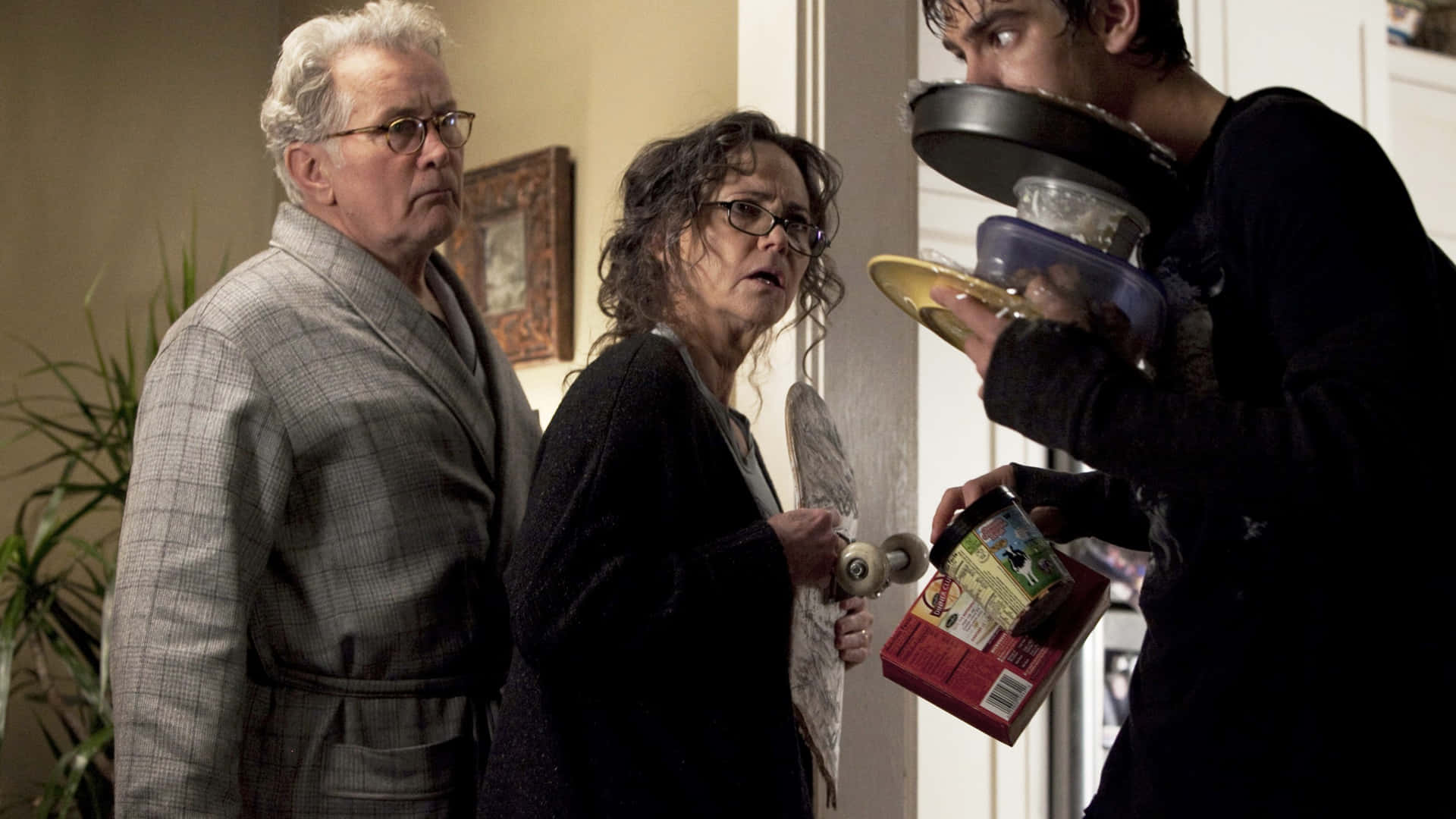Aunt May 桌布 壁紙 — 一對年長夫婦震驚地凝視著，一名身穿深色衣服的年輕男子拿著廚房用具和一個盒子，同時將煎鍋舉在臉上。